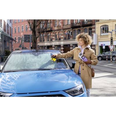 Een persoon in een beige jas ontdooit de voorruit van een blauwe auto met een elektrische ontdooier. Stadsgebouwen op de achtergrond.