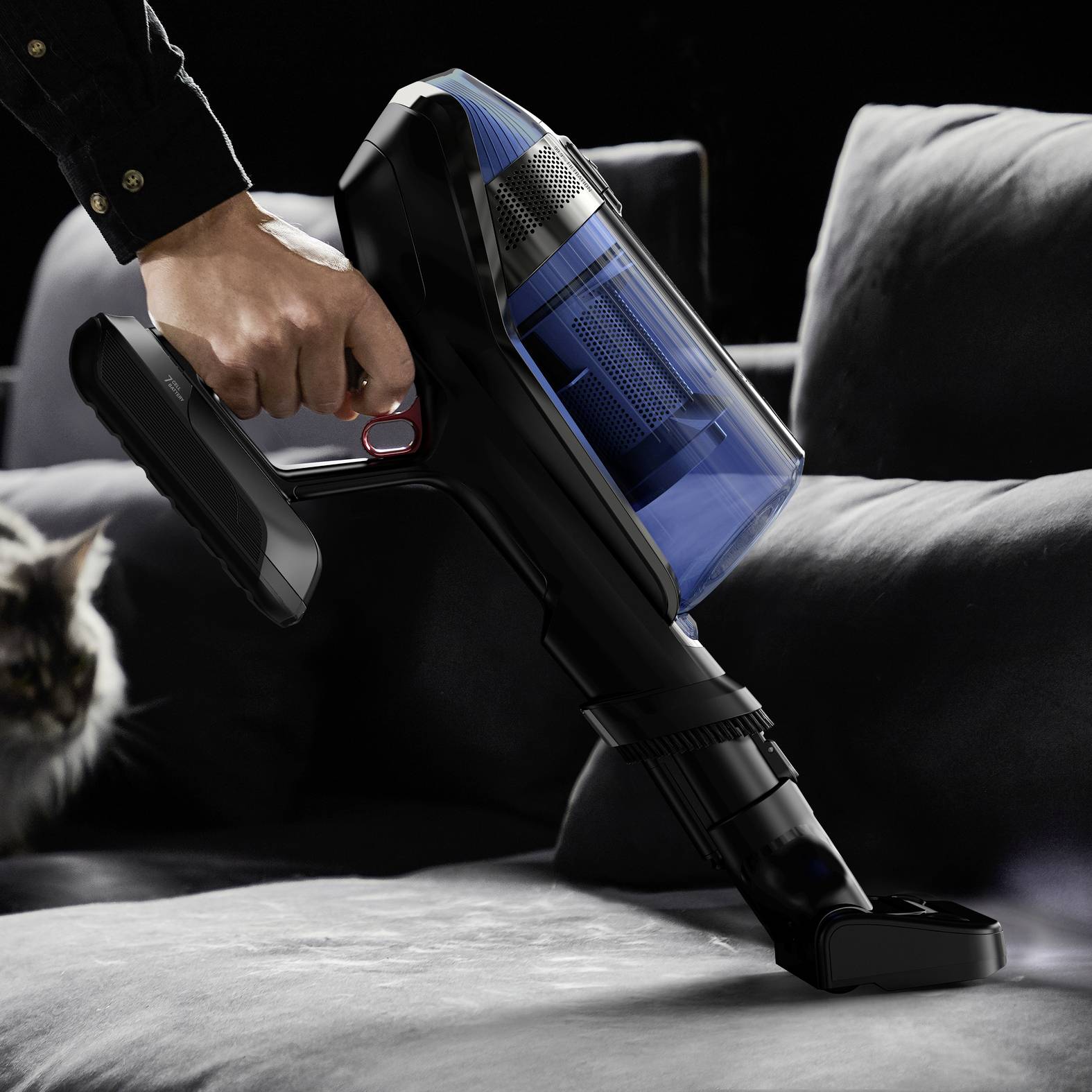 Een persoon zuigt een grijze bank schoon met een draadloze stofzuiger. Een kat zit op de achtergrond.