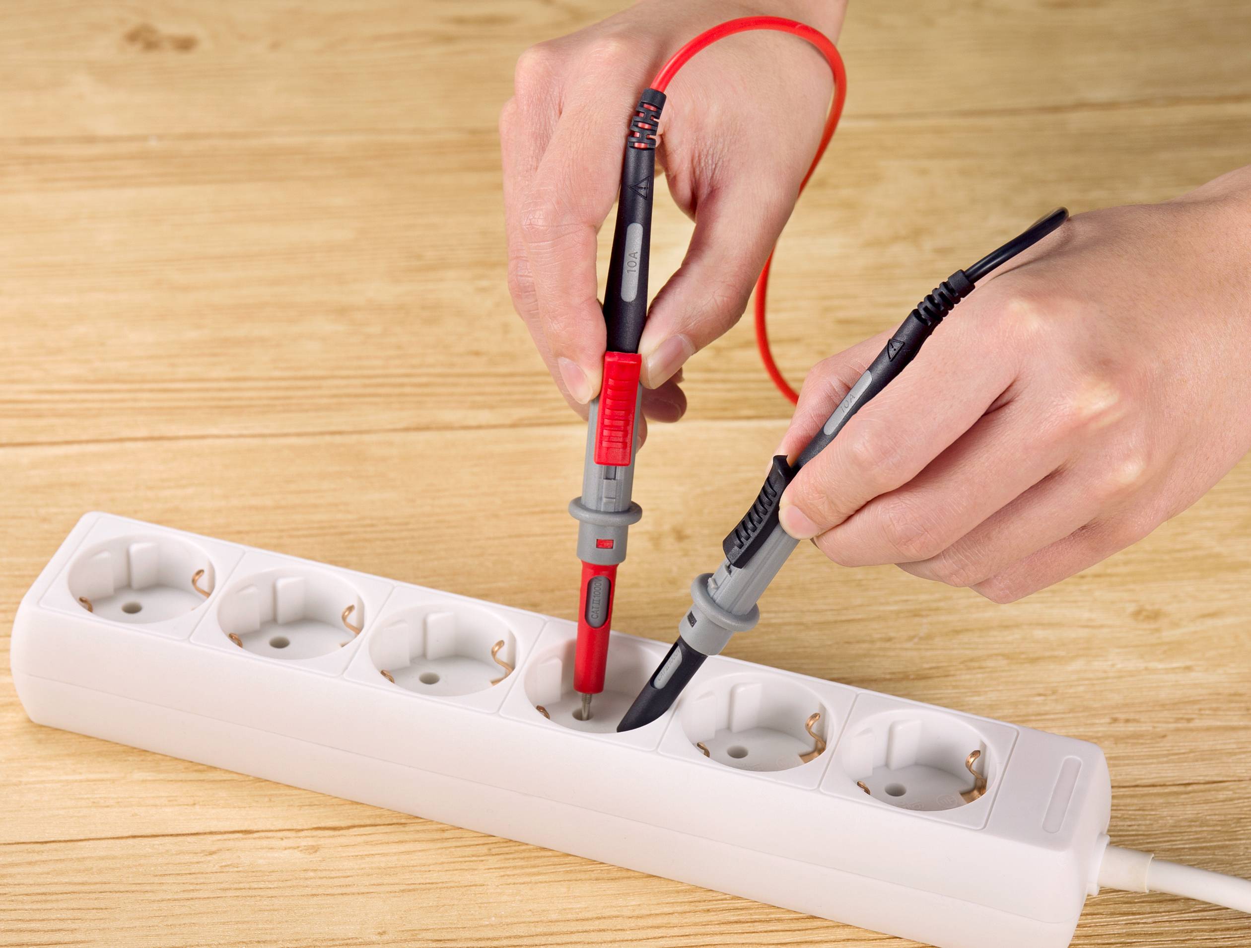 'Een persoon controleert met een multimeter de spanning van een witte stekkerdoos op een houten tafel.'