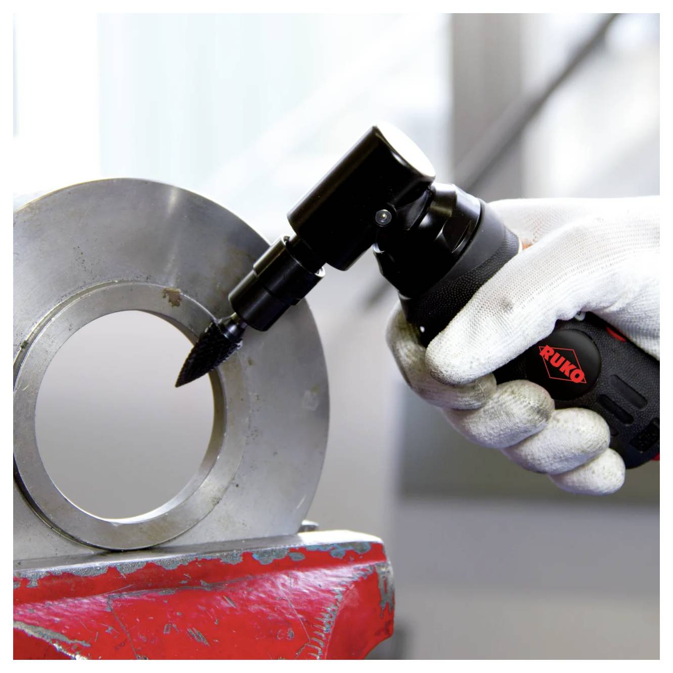 Gehandschoende hand die een zwart pneumatisch slijpgereedschap gebruikt op een grote metalen ring die in een rode bankschroef wordt vastgehouden.