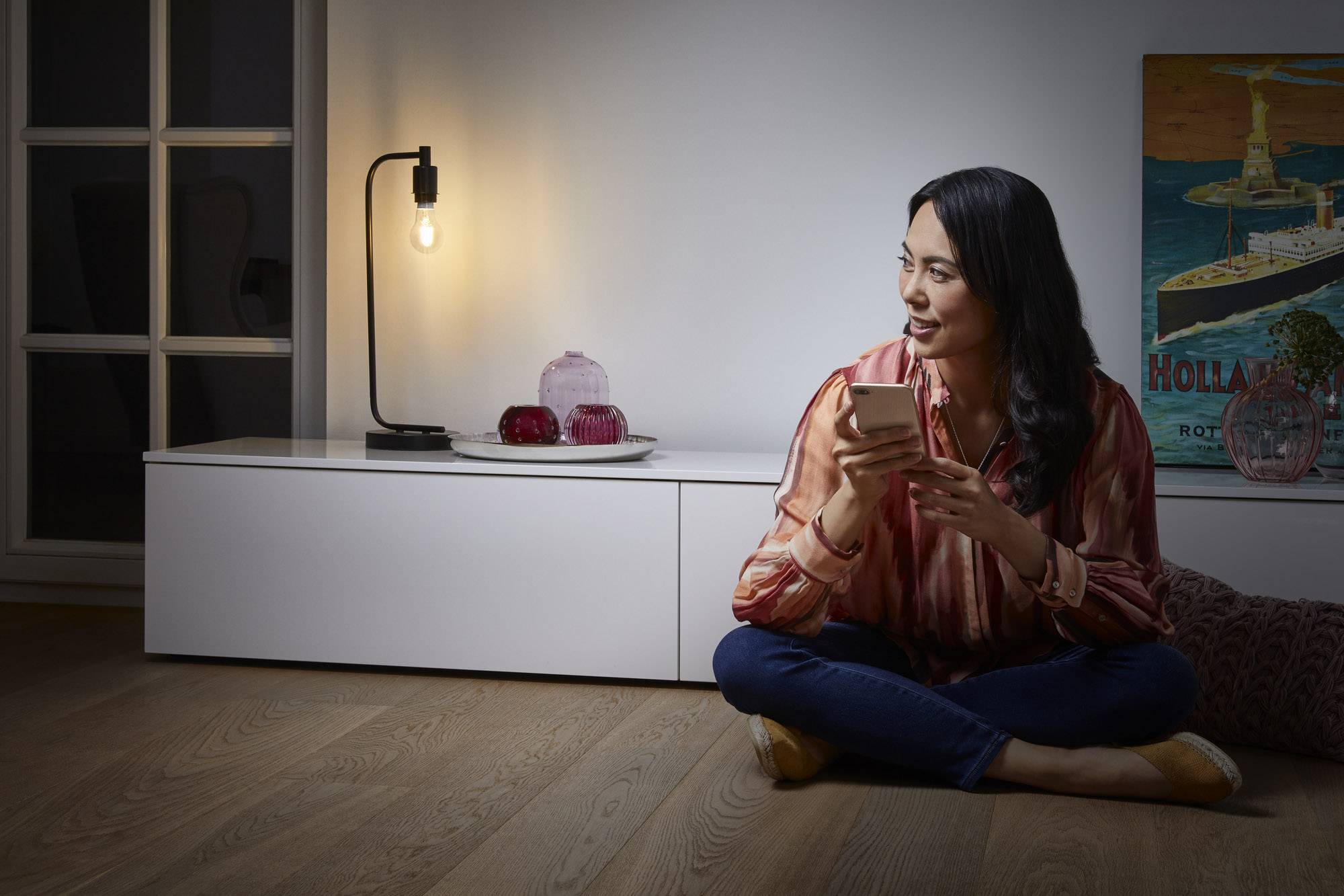 Een vrouw zit op de grond en houdt een smartphone vast, omringd door lamplicht en een wandposter op de achtergrond.