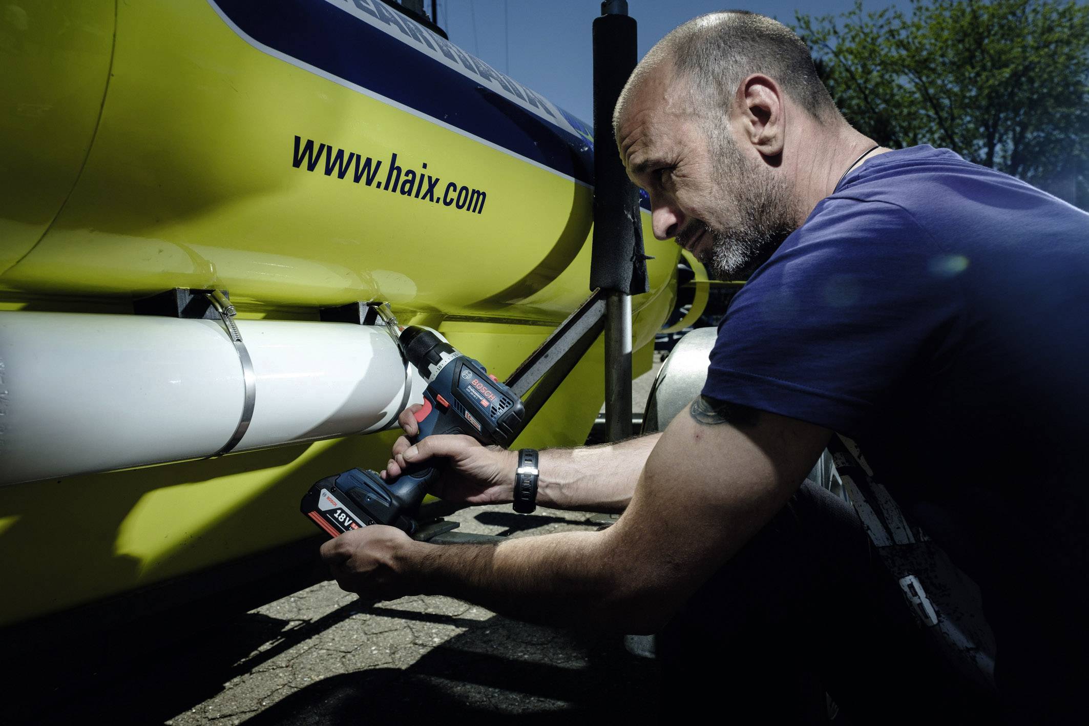 Een man gebruikt een accuschroevendraaier om iets te bevestigen aan een gele boot. Op de boot is 'www.haix.com' te zien.