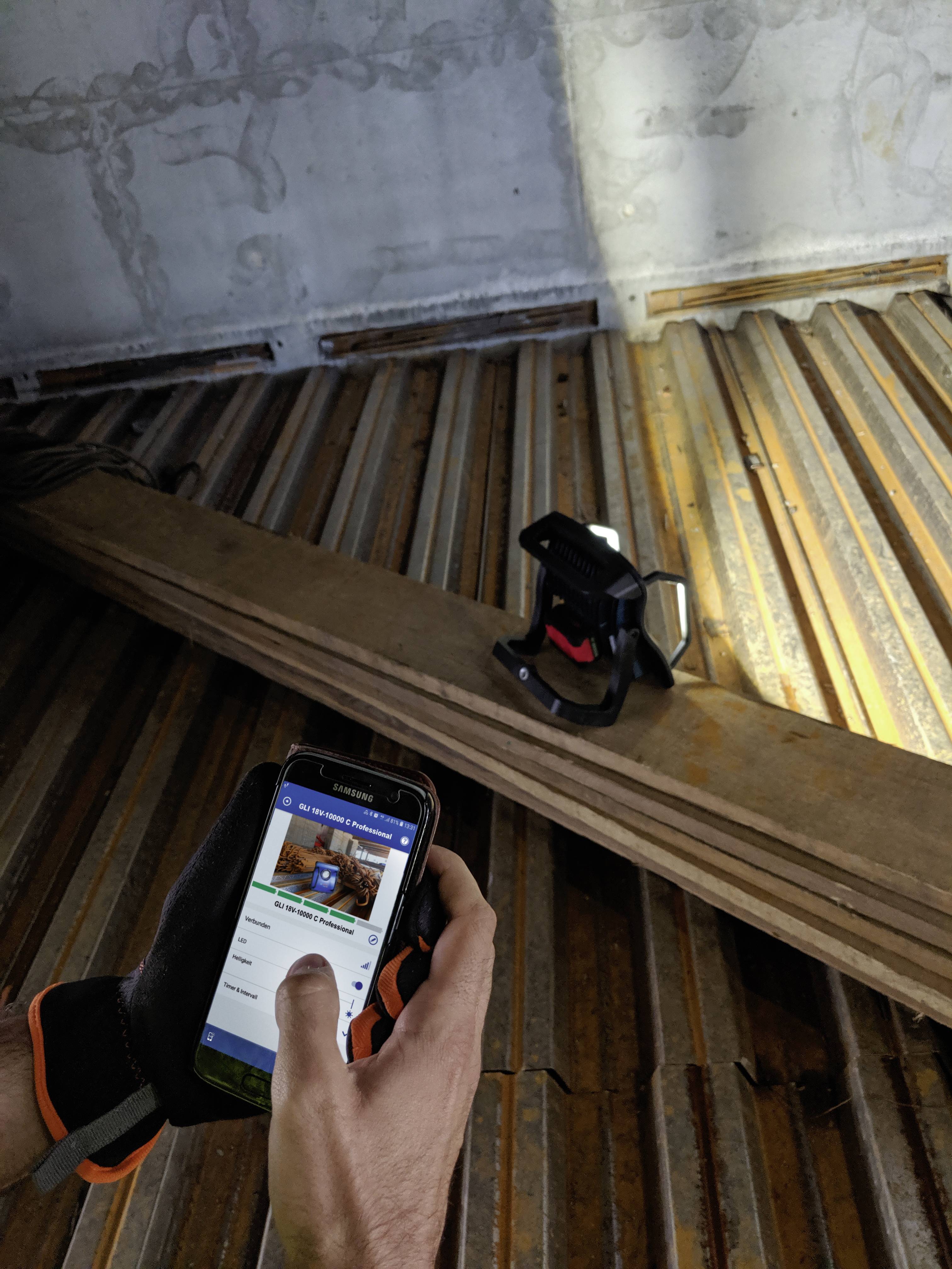 Een persoon met handschoenen bedient een mobiele telefoon terwijl hij met een lamp schijnt op een metalen oppervlak, waarschijnlijk voor inspecties.