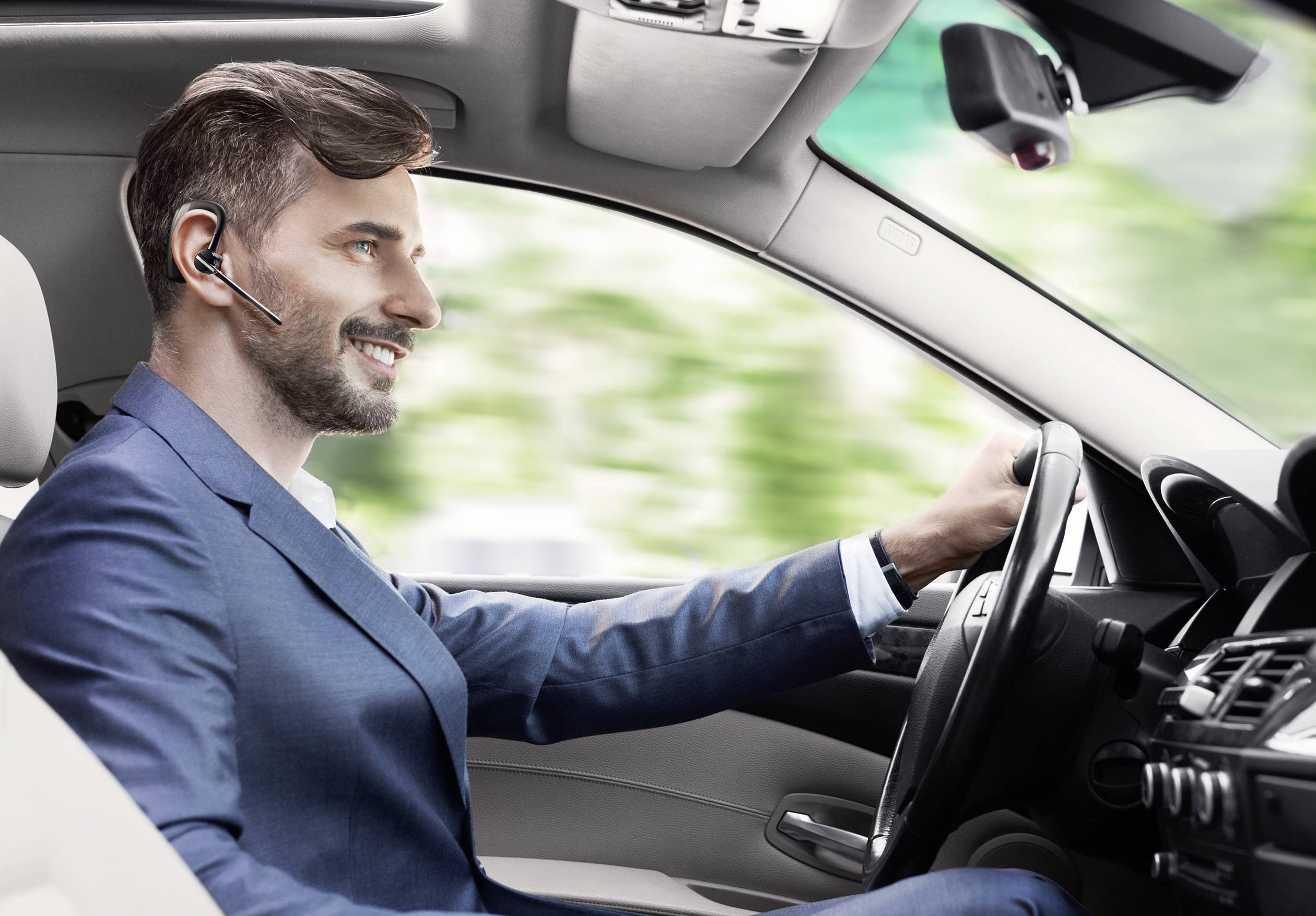 Een man in een pak rijdt een auto en spreekt via een headset. Op de achtergrond zijn wazig groene en witte kleuren zichtbaar.