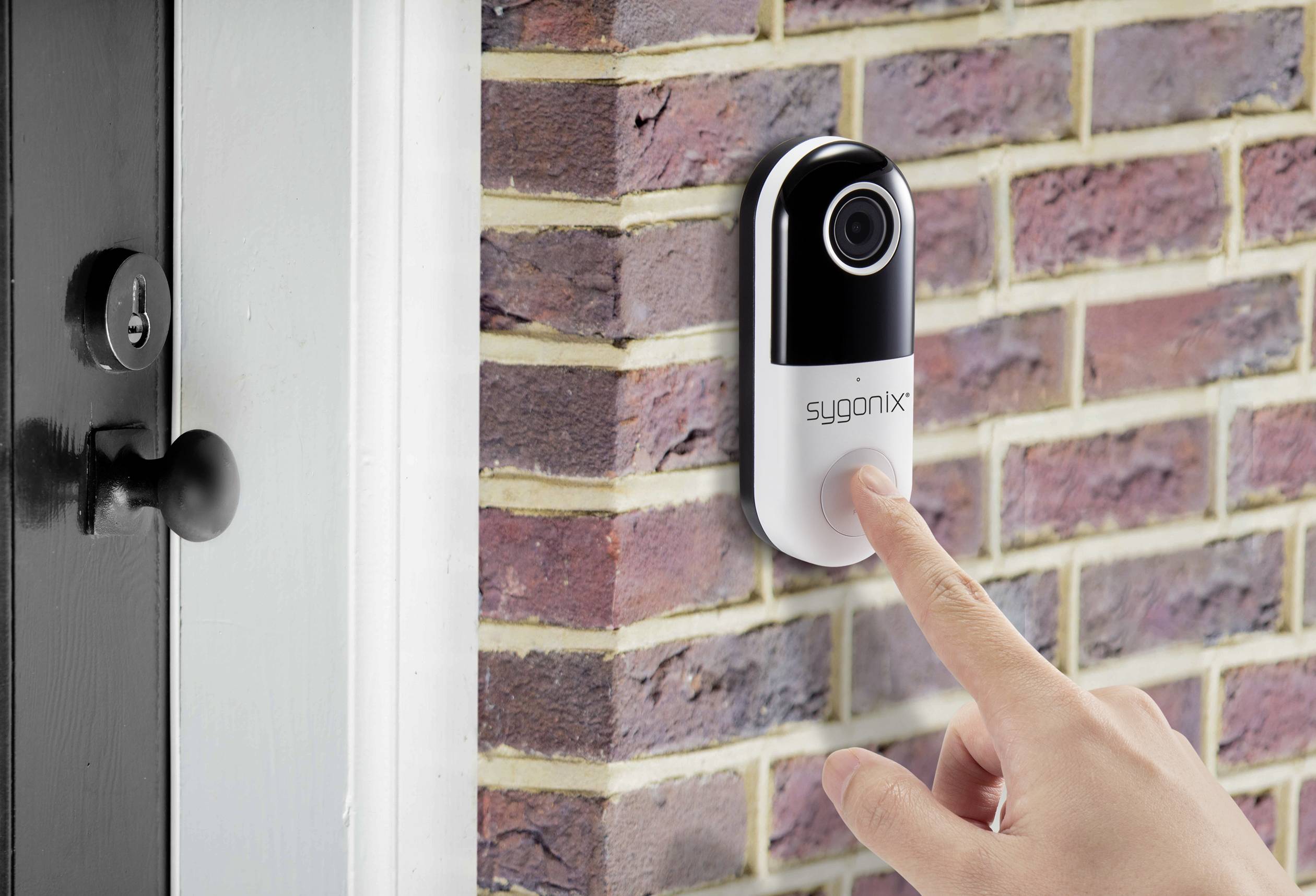 Een hand drukt op een deurbel met camera, die aan een bakstenen muur naast een zwarte deur is bevestigd.