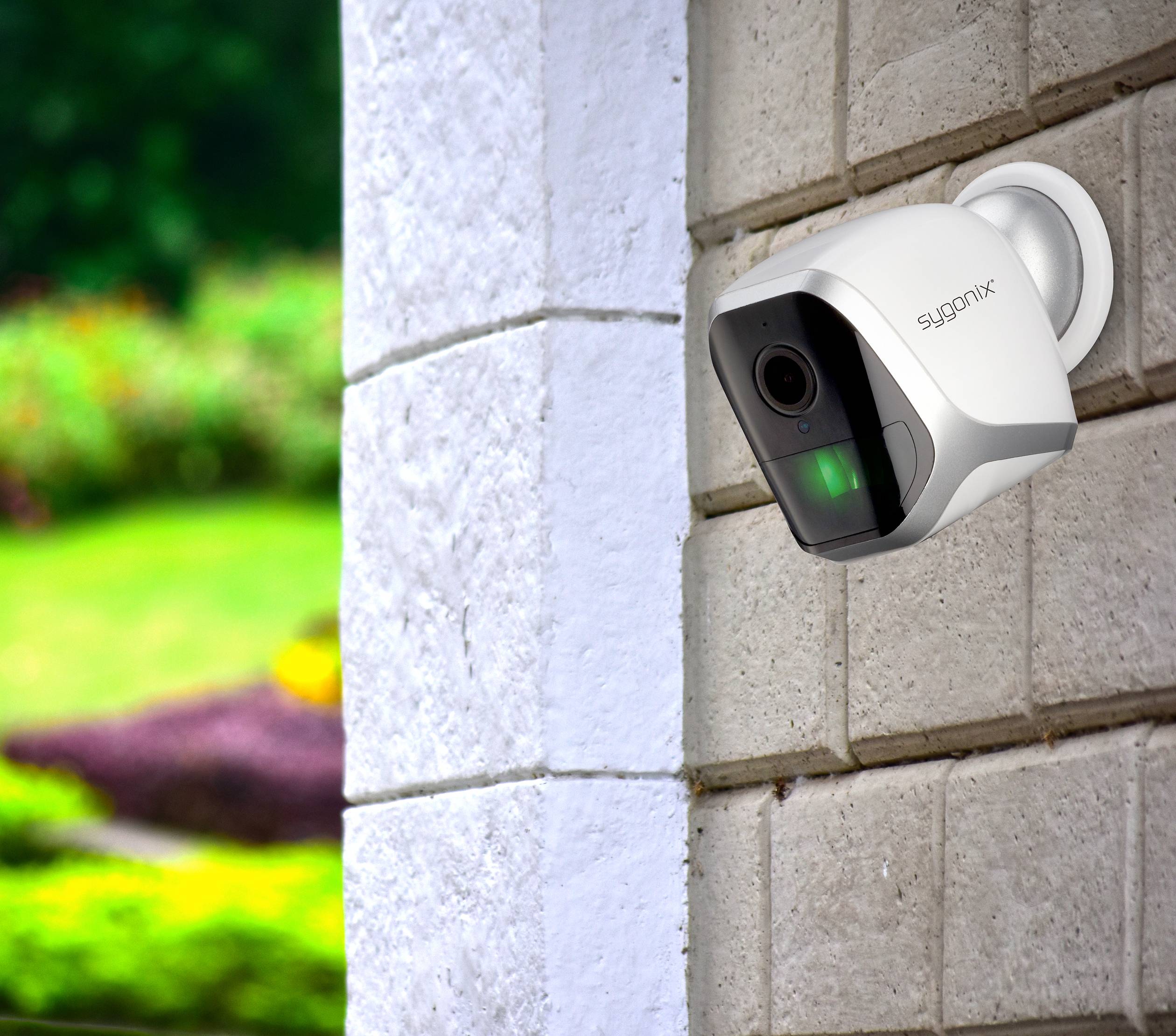 Een bewakingscamera is bevestigd aan een bakstenen muur en houdt toezicht op een groene tuin op de achtergrond.