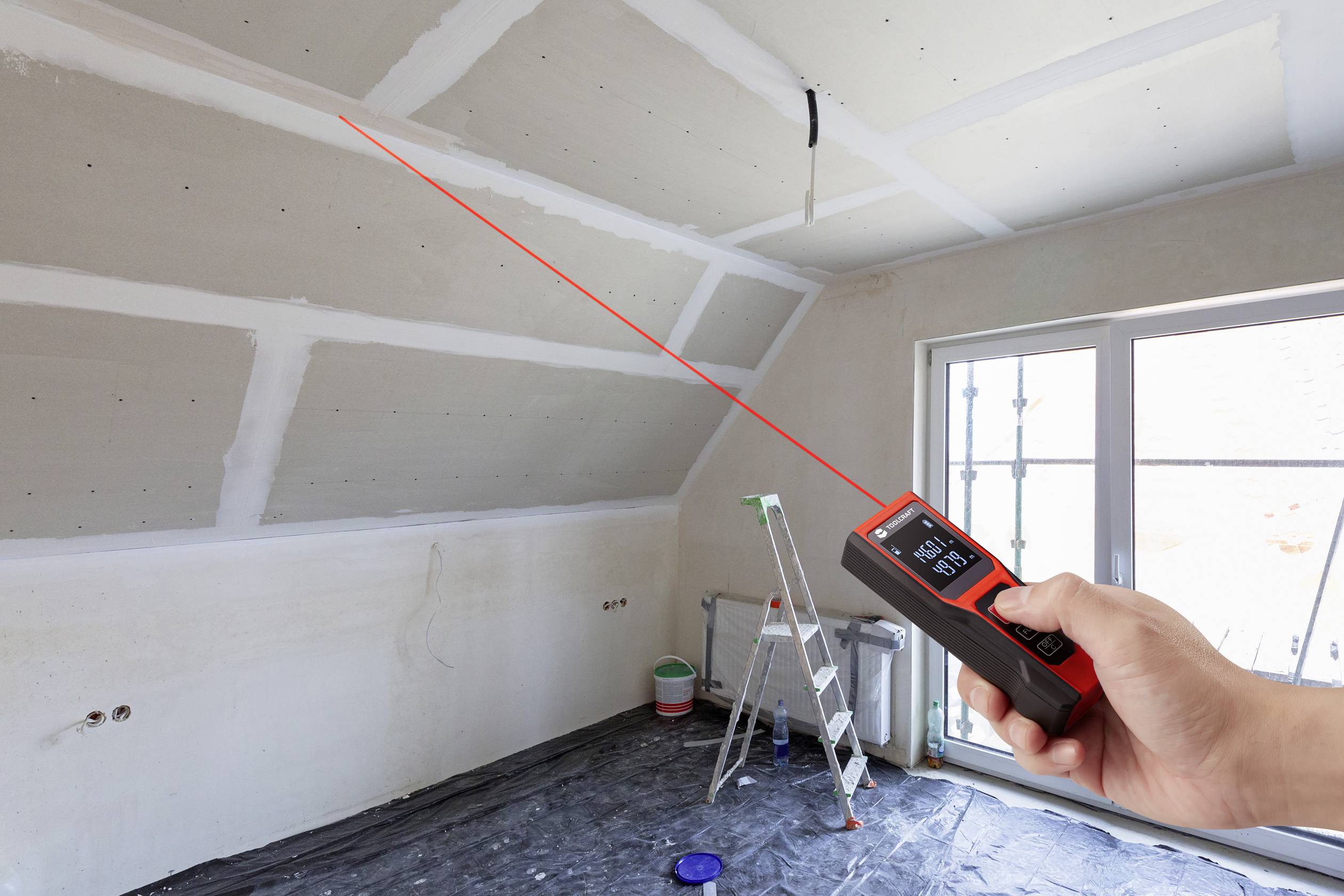 Een hand houdt een laserafstandsmeter vast in een ruimte in aanbouw, muren zijn klaar voor het schilderen, een ladder en verfpot op de achtergrond.