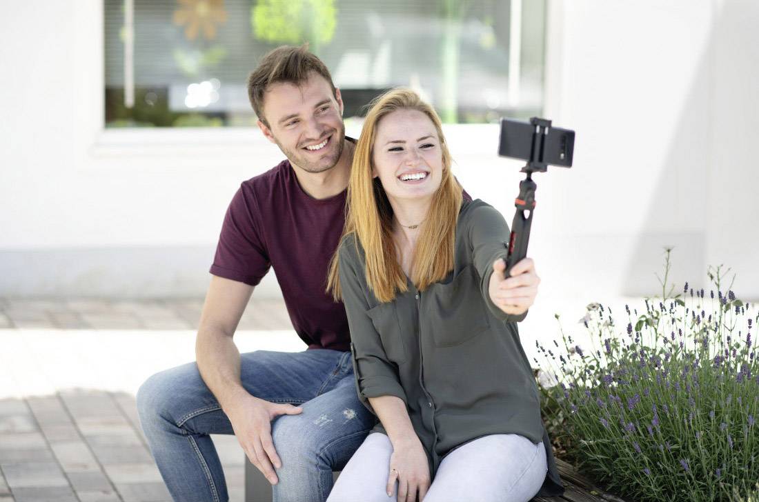 Een vrouw en een man zitten buiten op een bank en glimlachen naar de camera terwijl ze een selfie maken met een smartphone.
