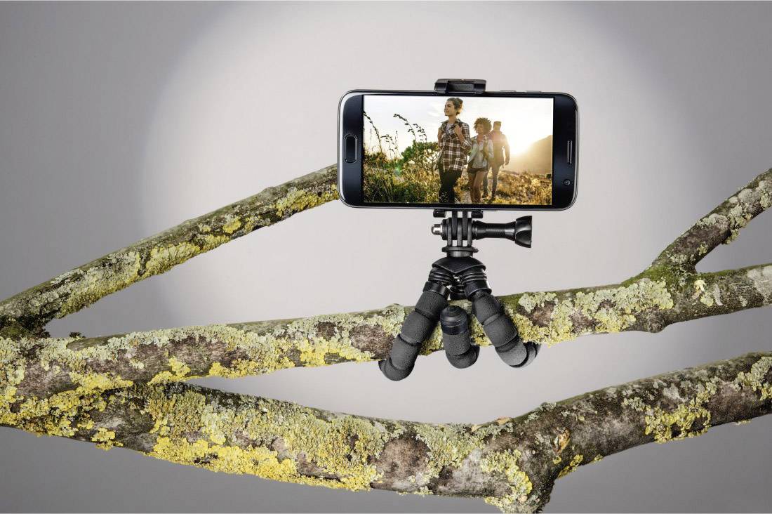 Een smartphone op een flexibele statief, bevestigd aan een tak, toont een afbeelding van twee personen die wandelen in de natuur.