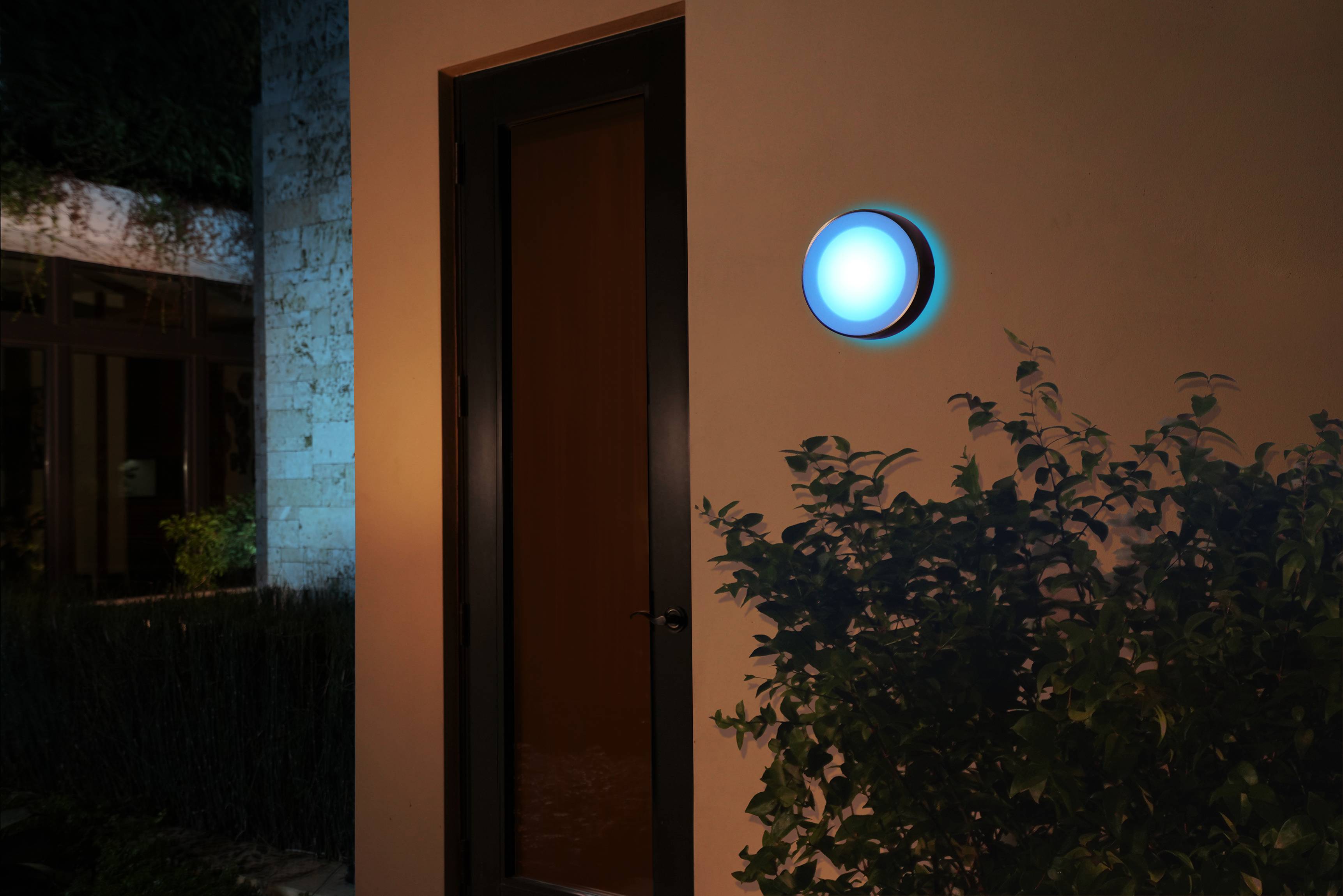 Buitenaanzicht van een huis 's nachts met een rond, helder blauw licht aan de muur naast een deur, omgeven door planten.