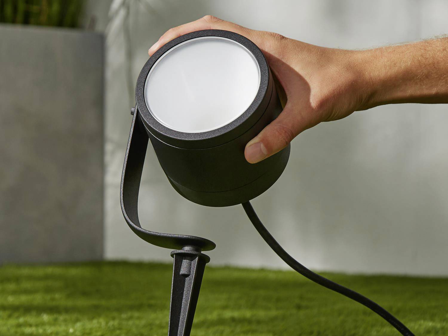 Een hand richt een zwarte buitenspot uit op groen gras. De focus ligt op de spot en de handbeweging.