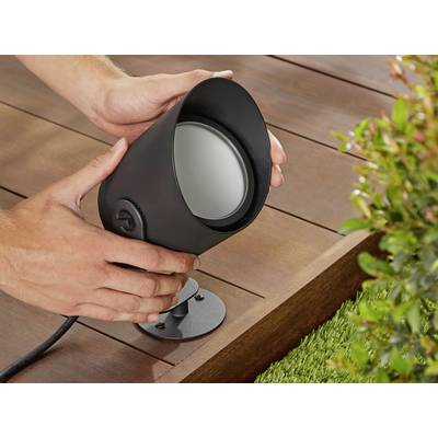 Een persoon installeert een zwarte buitenlamp op een houten terras, nabij een gebied met groene planten.