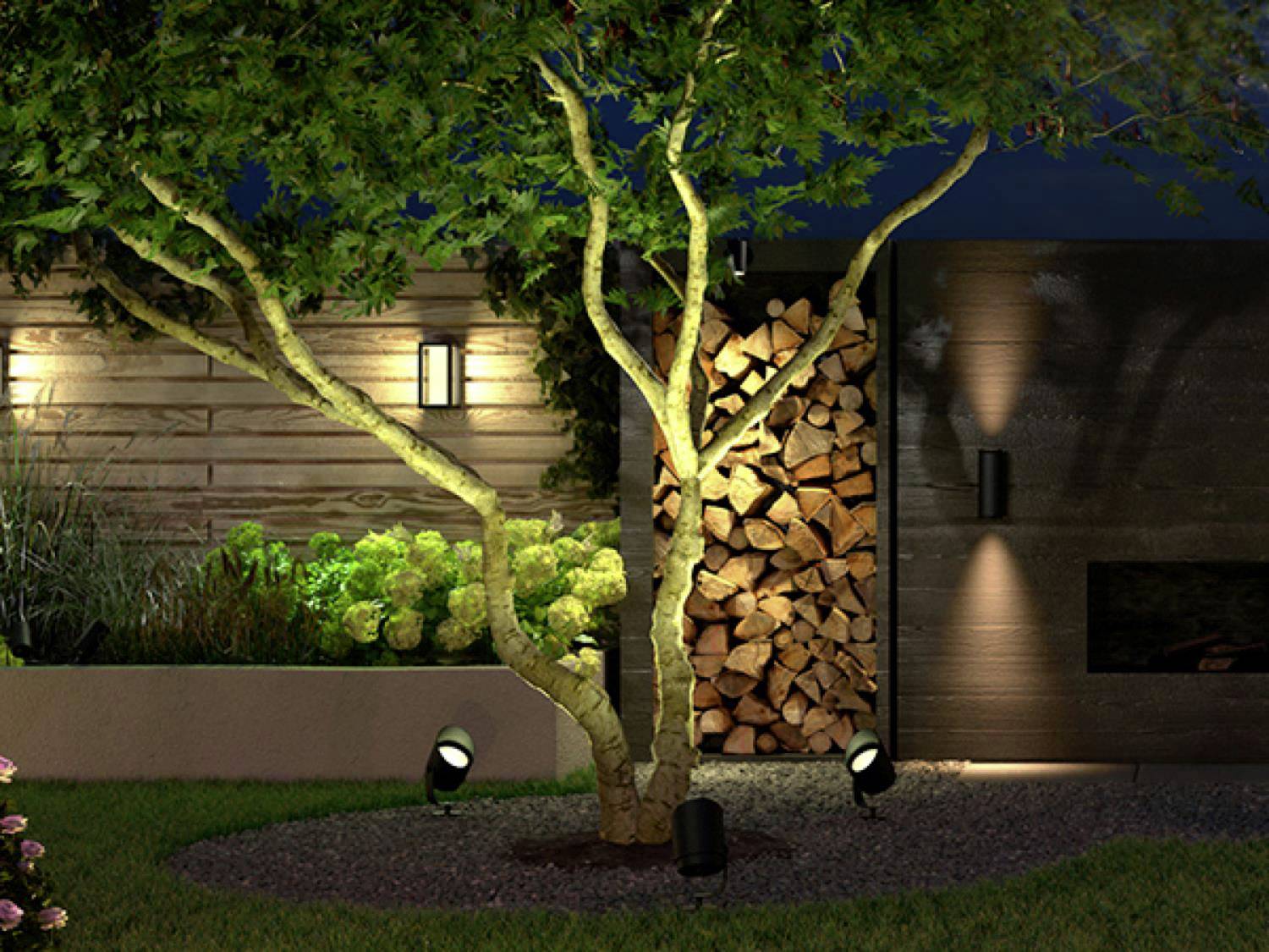 Een verlichte tuin met een boom in het centrum, omringd door schijnwerpers. Op de achtergrond zijn stapels hout en een houten wand zichtbaar.