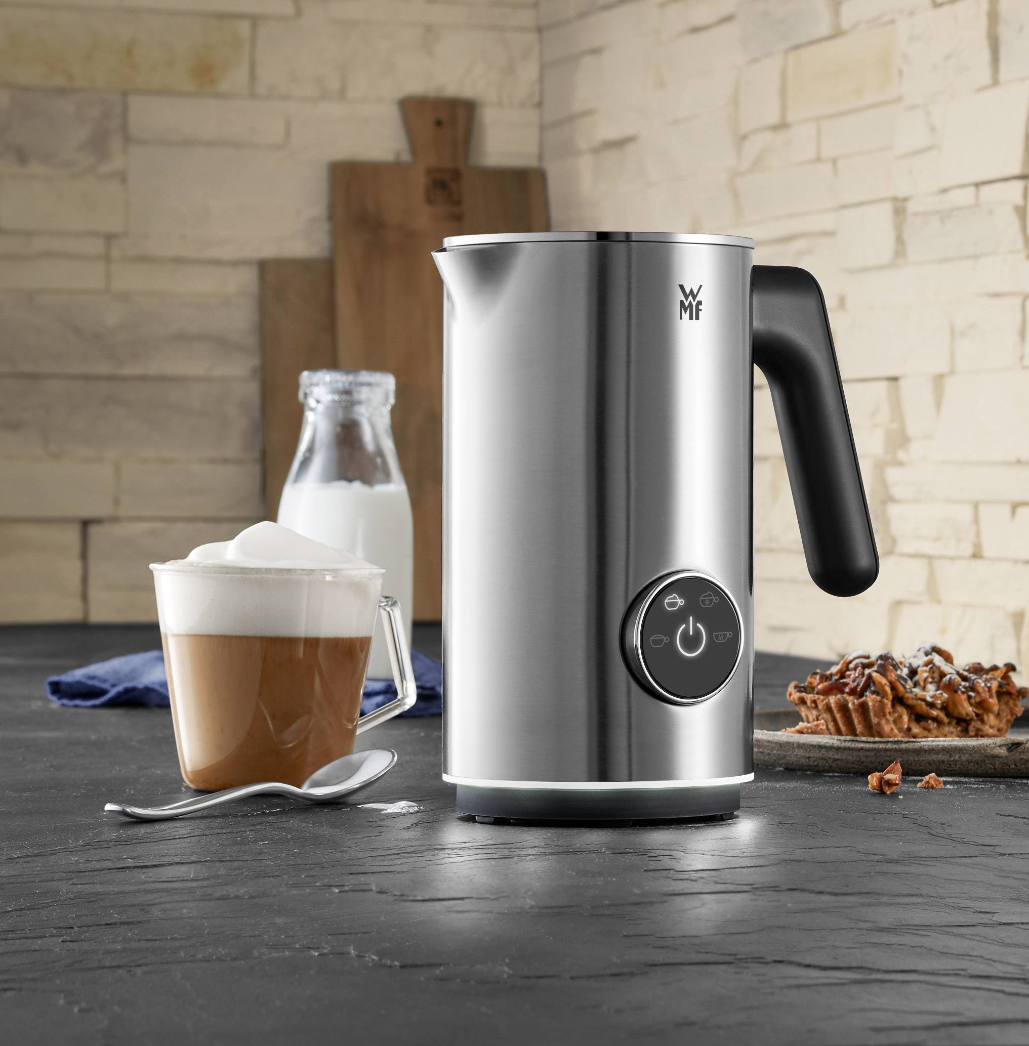 Een elektrische melkopschuimer van roestvrij staal staat op een aanrecht. Ernaast een kopje cappuccino en een glas melk.
