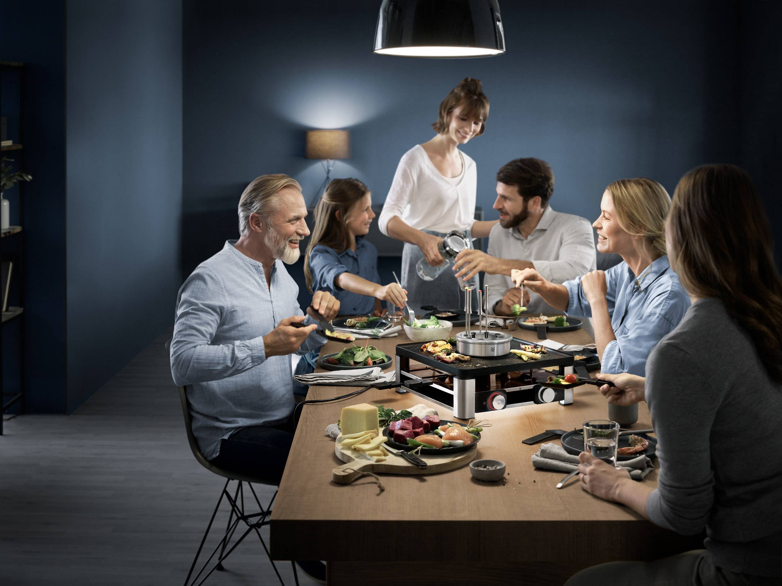 Een groep van zes personen zit glimlachend rond een tafel en geniet van een gezamenlijke maaltijd in een gezellige, donkere omgeving.