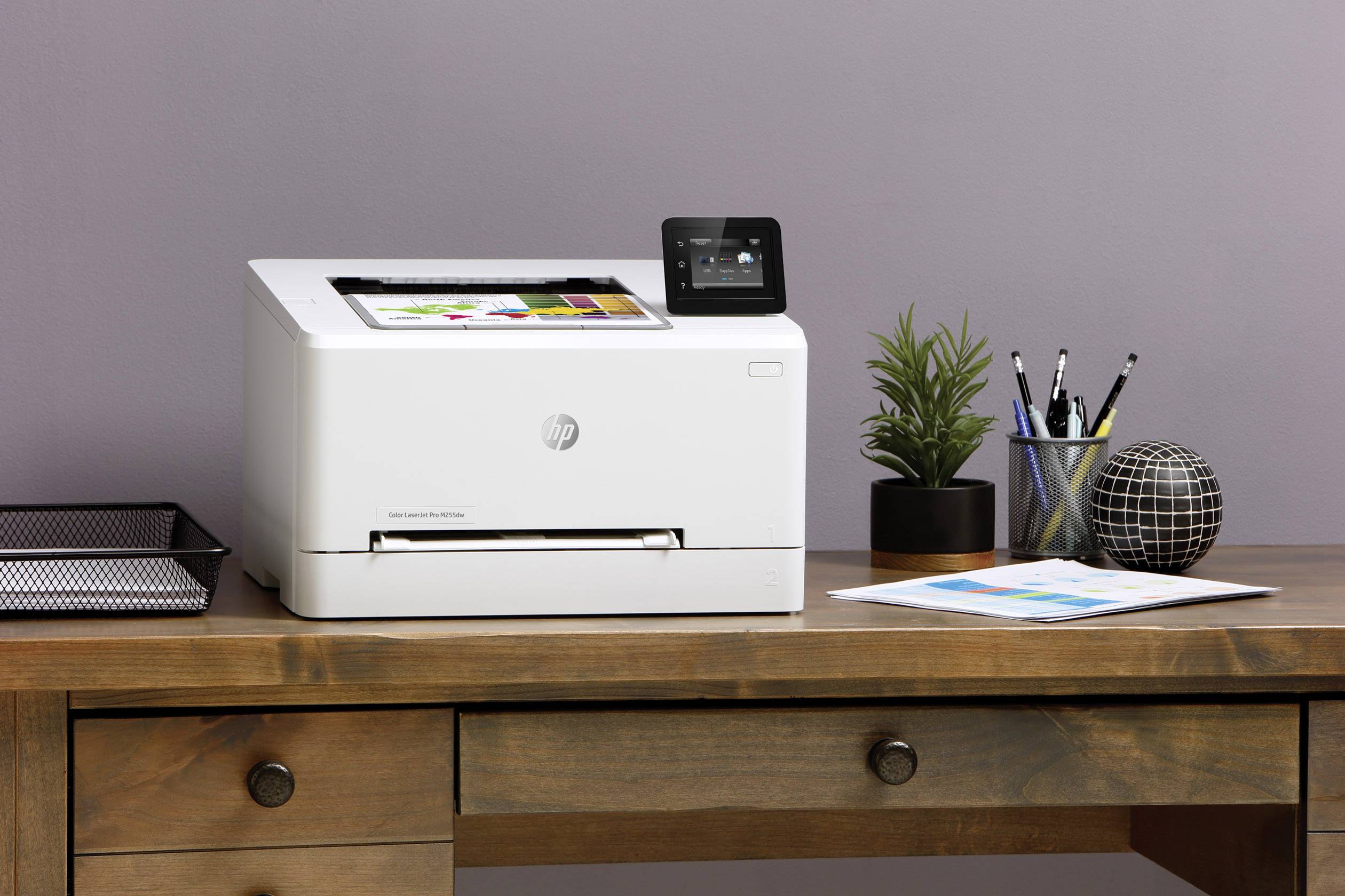 Witte printer op een houten tafel, naast planten, schrijfwaren en papier, tegen een paarse muur.