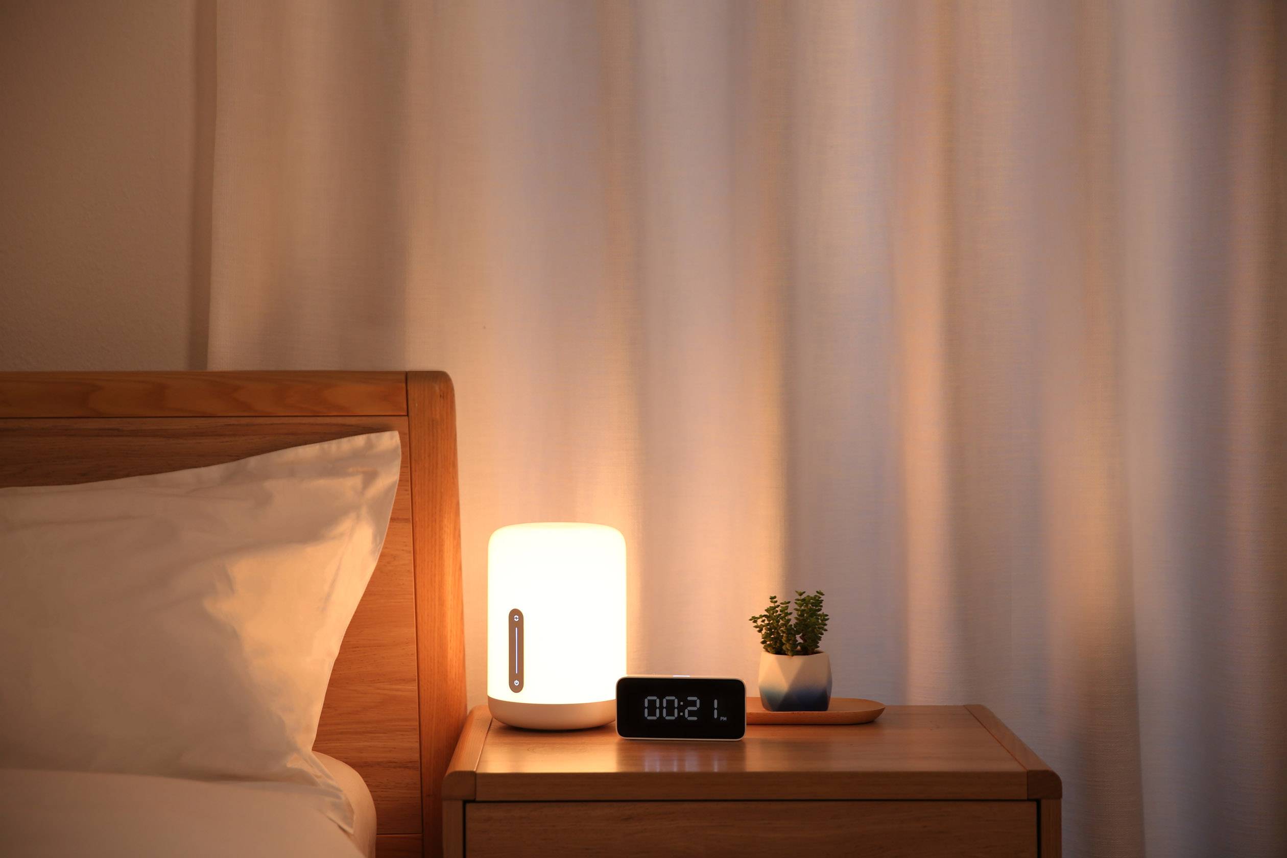 Een nachtkastje met een verlichte lamp, een digitale wekker die '00:21' aangeeft, en een kleine plant erop.
