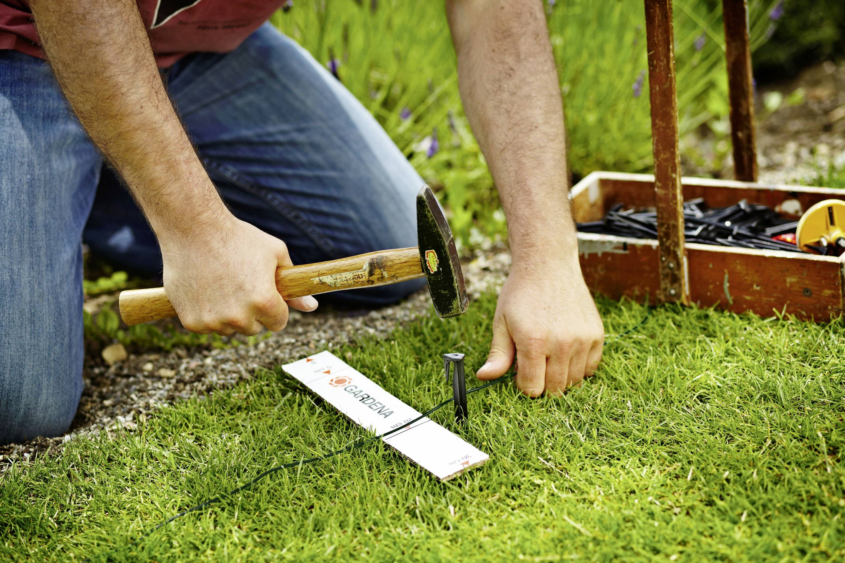 Een persoon knielt in het gras en slaat met een hamer een spijker in de grond. Op de achtergrond staat een gereedschapskist.