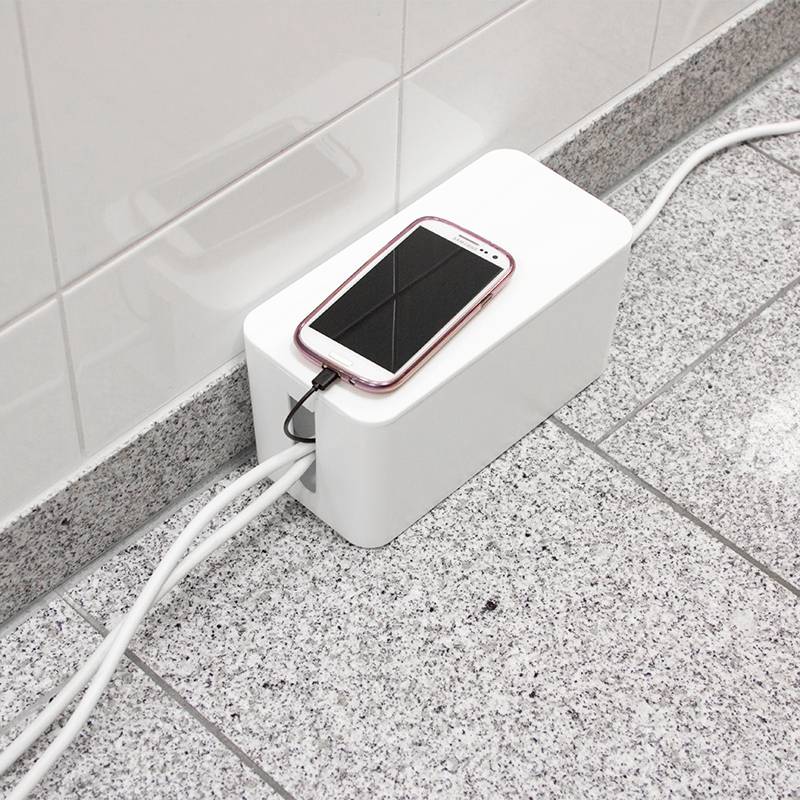 Een smartphone ligt op een witte kabeldoos op de grond. Verschillende kabels zijn naar de doos geleid om rommeligheid te voorkomen.