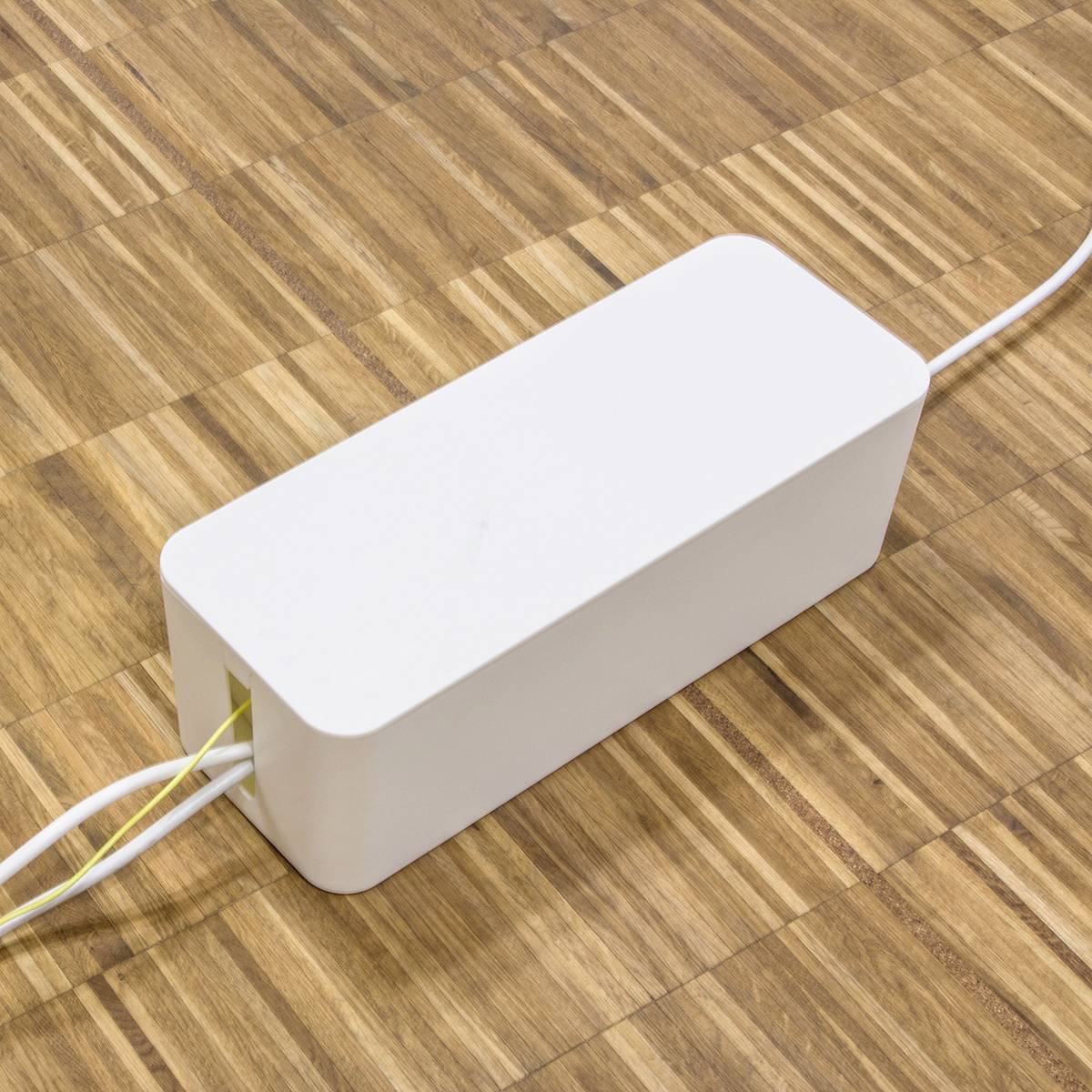 Een witte rechthoekige doos met kabels ligt op een houten vloer. Het zou kunnen gaan om een elektronisch apparaat of een adapter.