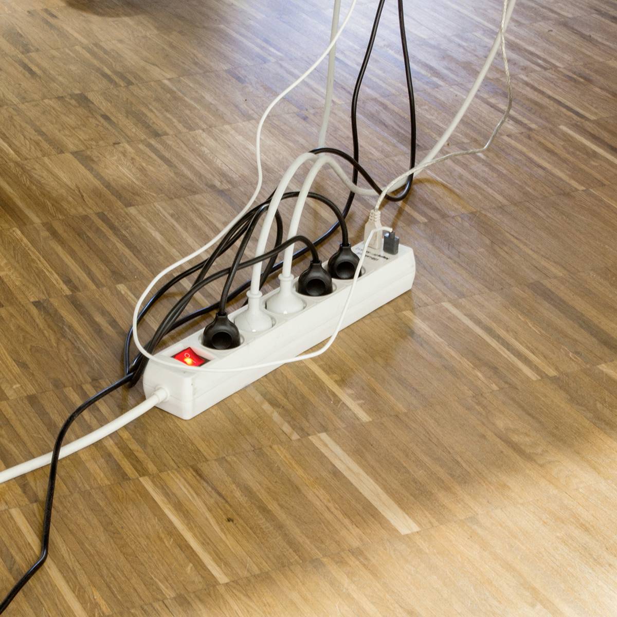 Meerdere elektrische apparaten zijn aangesloten op een stekkerdoos op een houten vloer. Een schakelaar toont rood, wat wijst op "ingeschakeld".