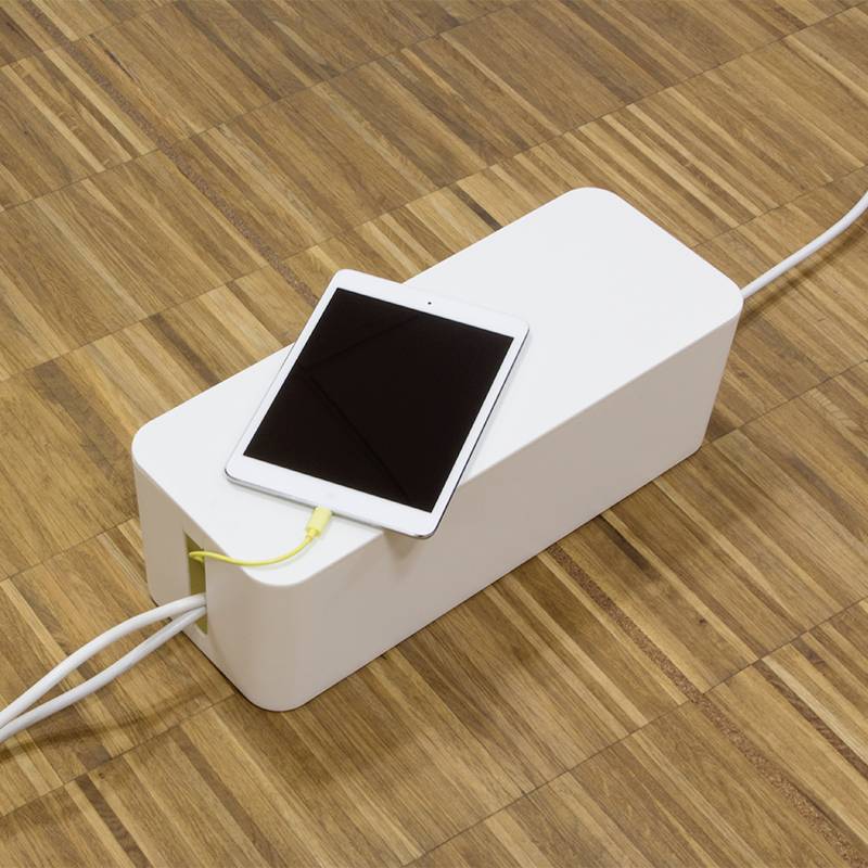 Een tablet ligt op een witte houten kabelbox op een houten vloer. Een oplaadkabel verbindt het tablet met de box.