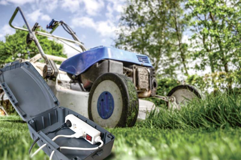 Een blauwe grasmaaier staat op hoog gras in de tuin, op de voorgrond ligt een geopende beschermhoes met een wit verlengingssnoer.