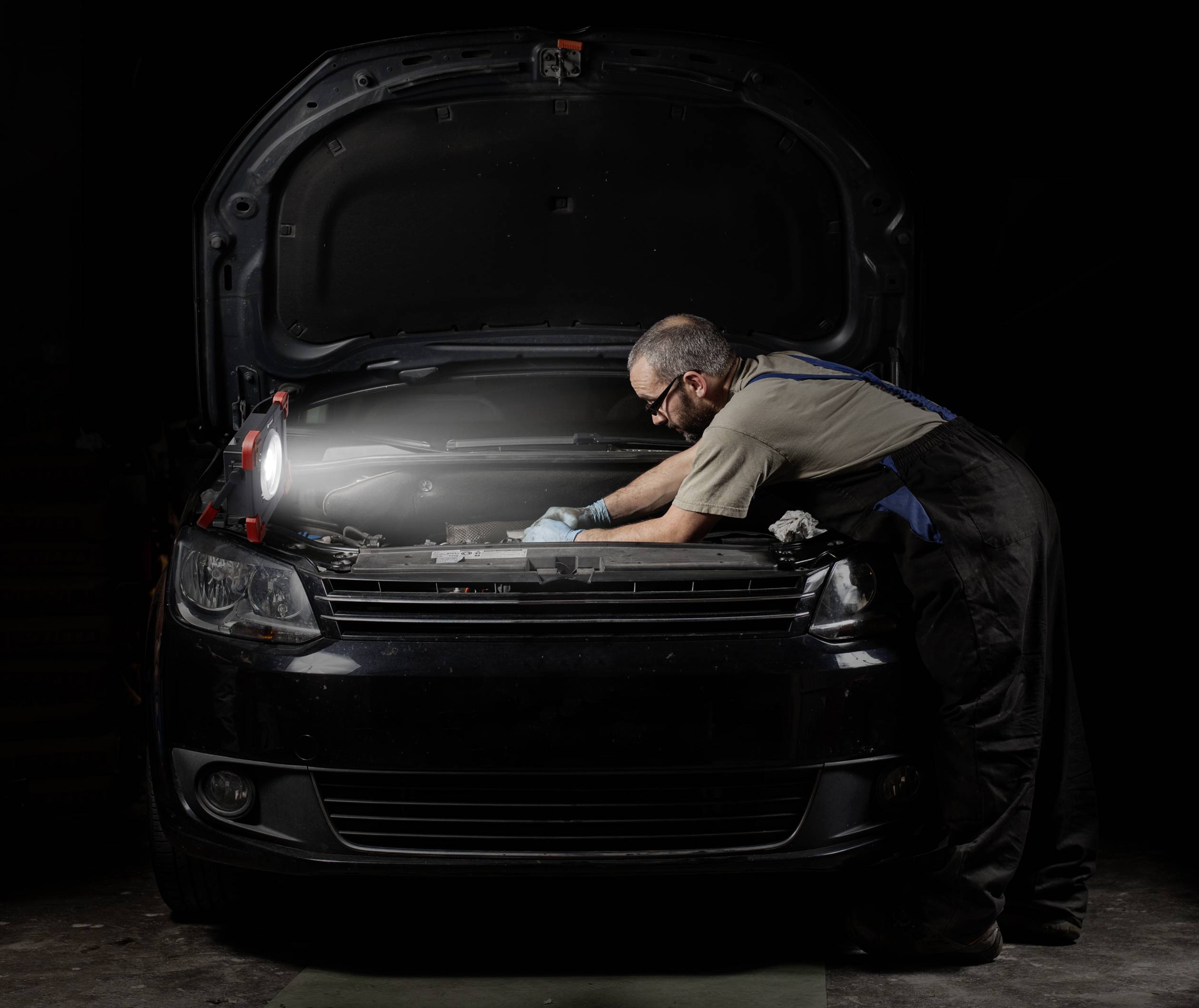 Een monteur buigt zich over de geopende motorkap van een auto en inspecteert de motor onder een felle lamp in een donkere omgeving.