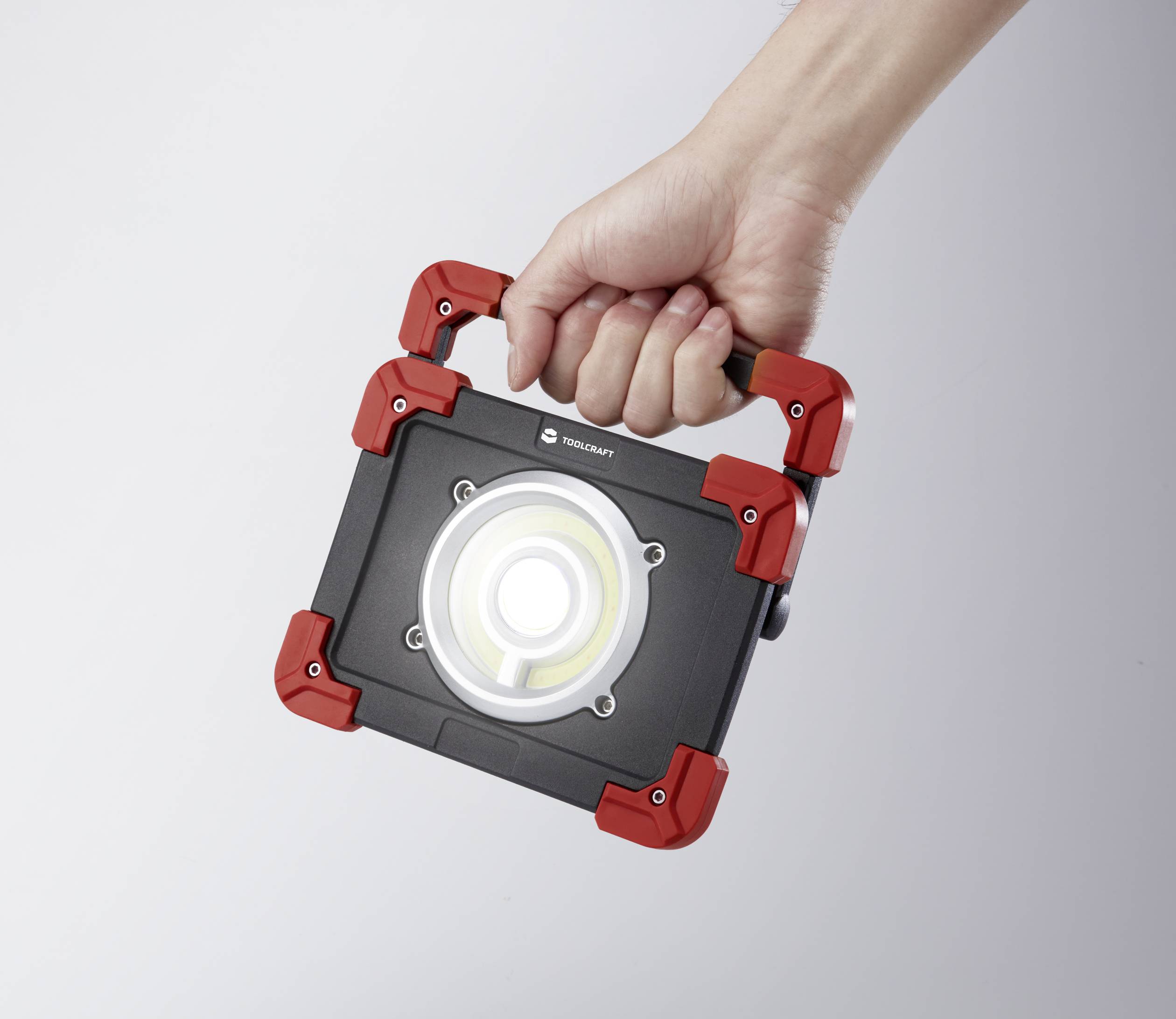 Een hand houdt een draagbare, rechthoekige LED-werkverlichting met een behuizing in rood en zwart.