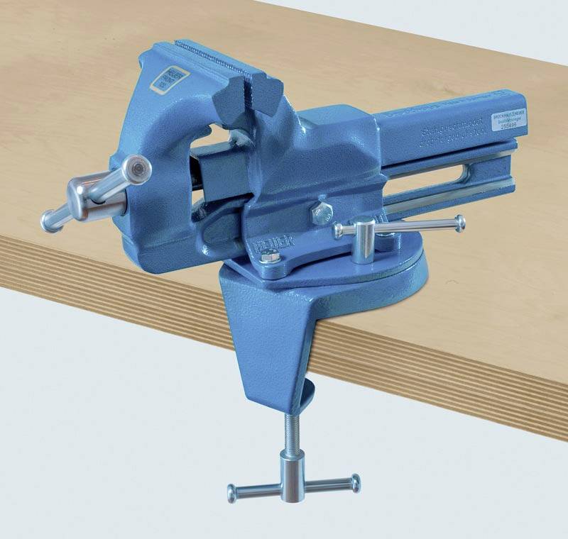Blauwe bankschroef bevestigd aan de rand van een houten tafel, dient ter stevige bevestiging van werkstukken tijdens ambachtelijke werkzaamheden.