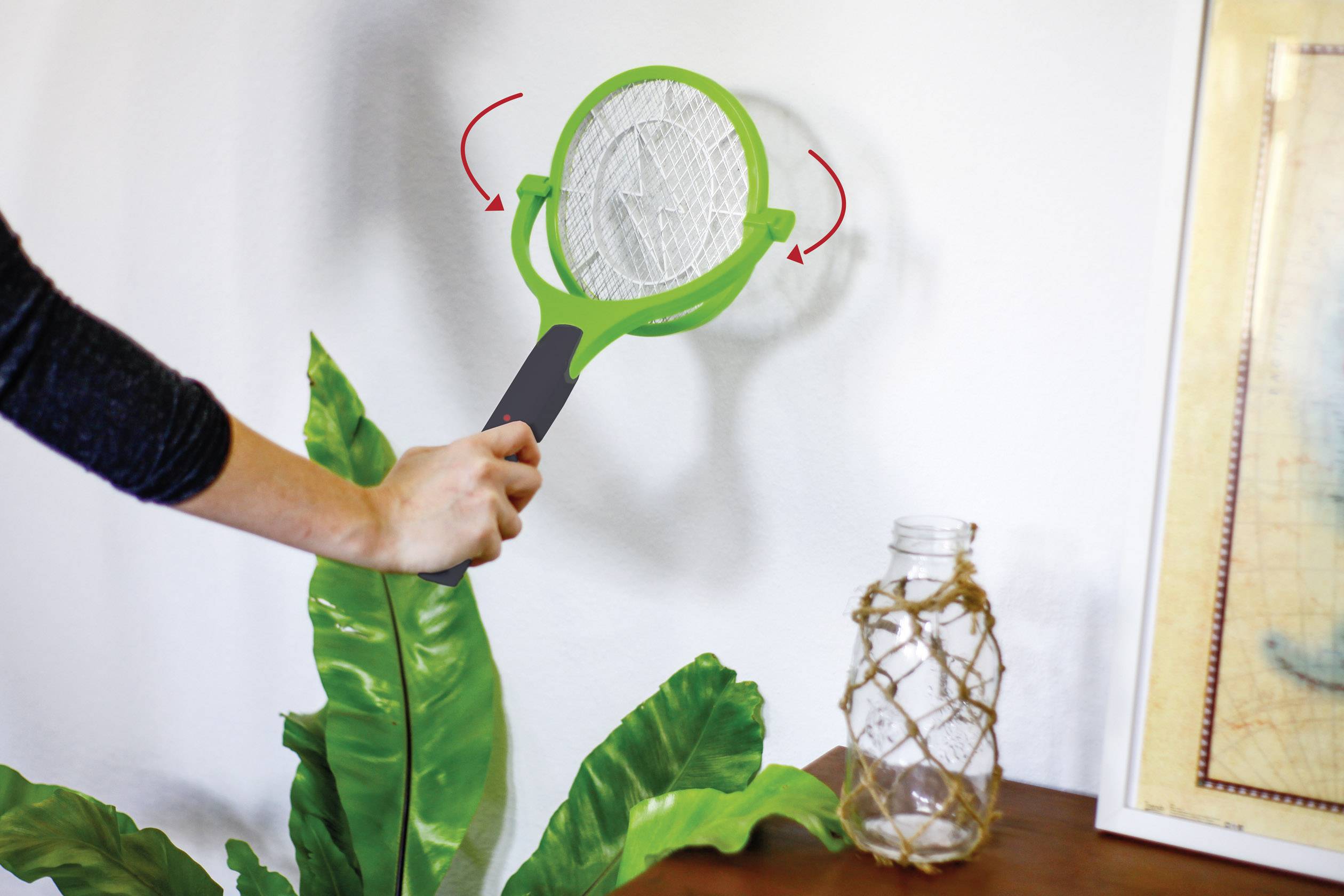 Een persoon houdt een elektrische vliegenmepper boven een plant om insecten te vangen. Inrichtingselementen zijn zichtbaar op de achtergrond.