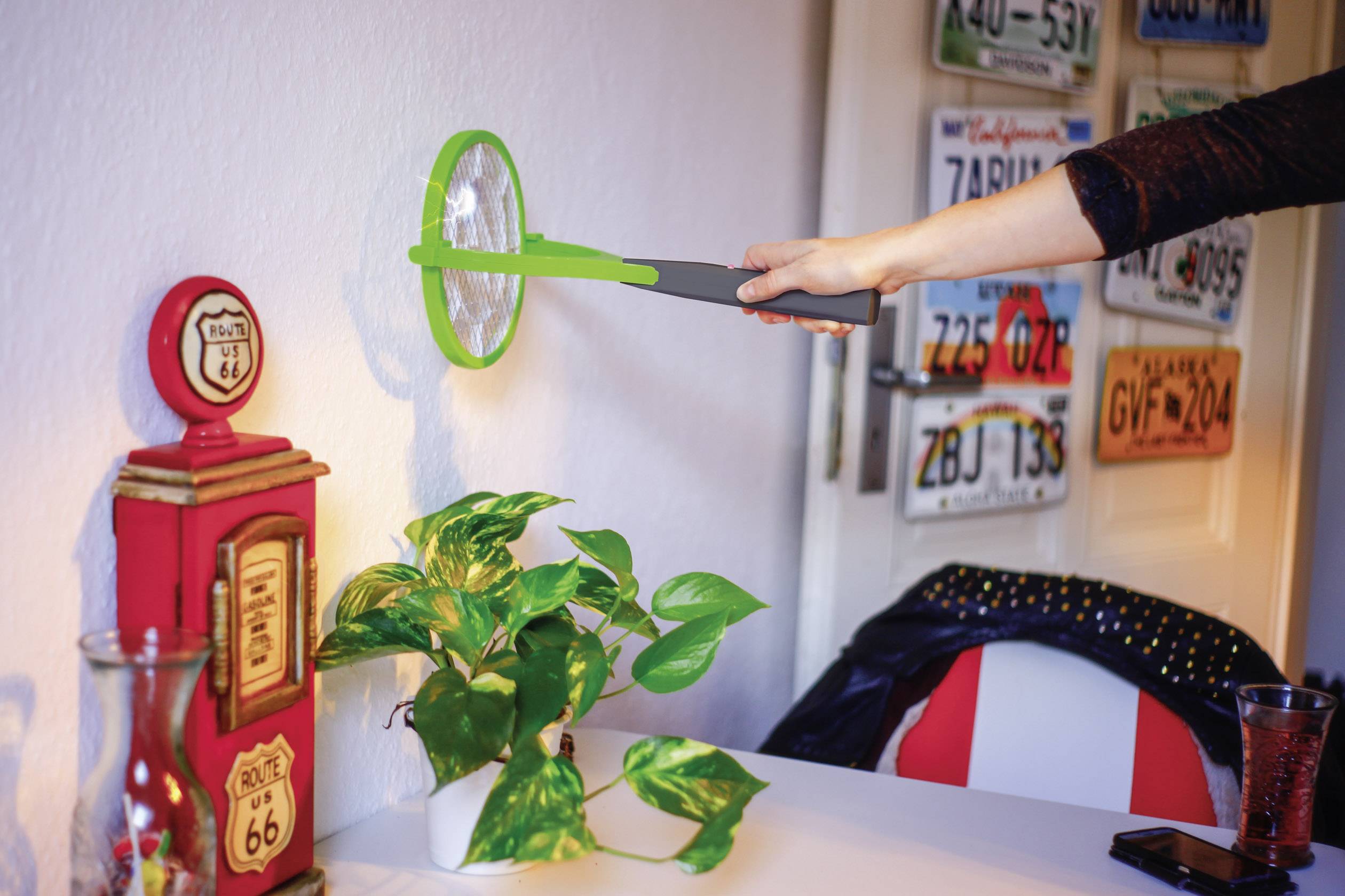 Een persoon slaat met een elektrische vliegenmepper op een groene plastic plant. Op de achtergrond zijn genummerde nummerplaten aan de muur.