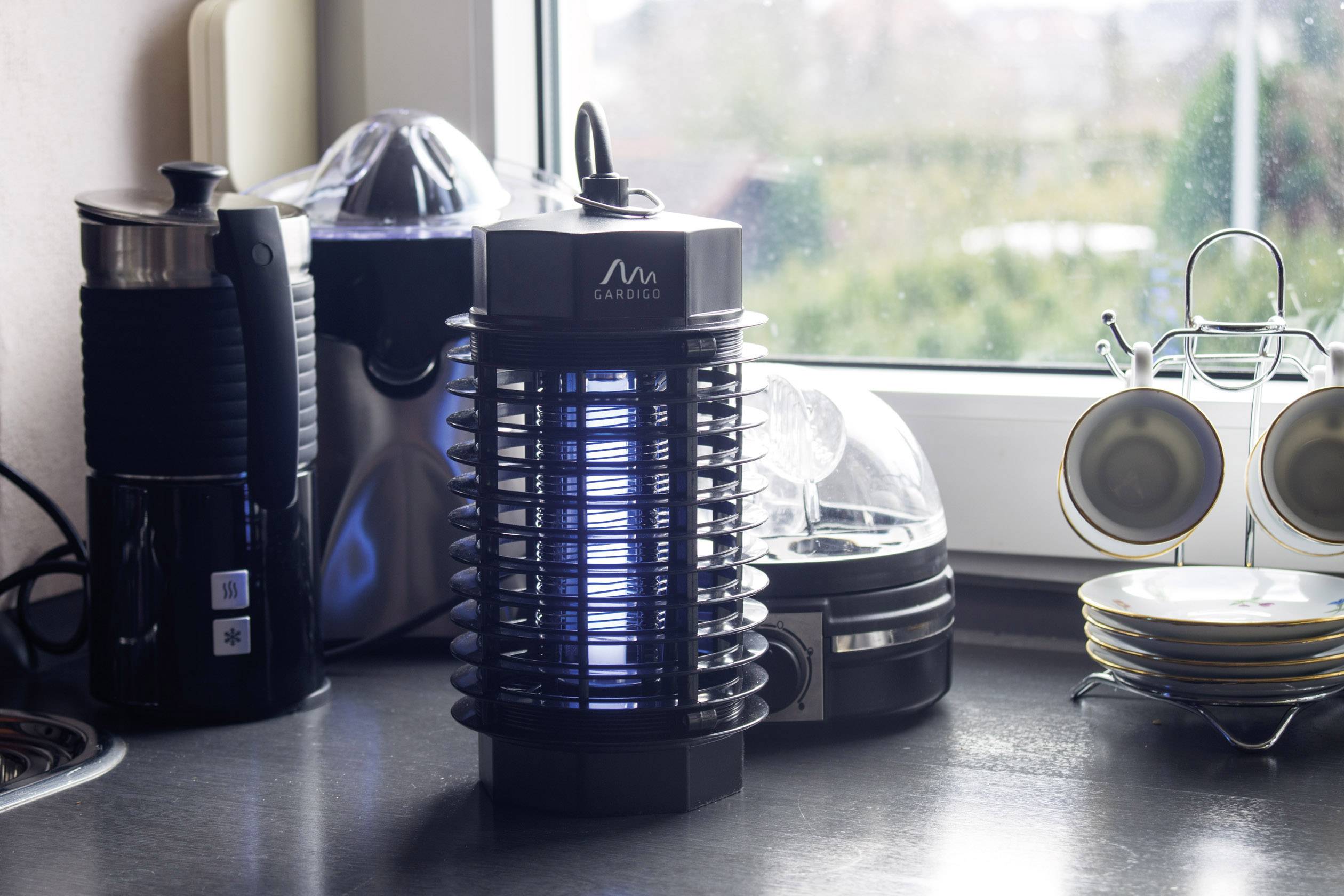 Elektrisch muggendodend apparaat op het aanrecht in een keuken, omgeven door servies en keukengerei, staat voor een raam.