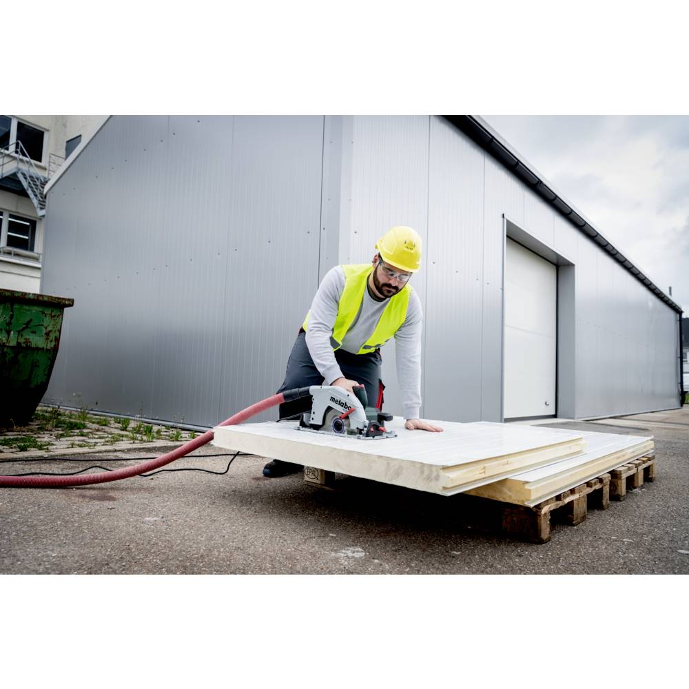 Een bouwvakker met een gele helm en veiligheidshesje zaagt witte platen door met een cirkelzaag, gepositioneerd op pallets in de buitenlucht.