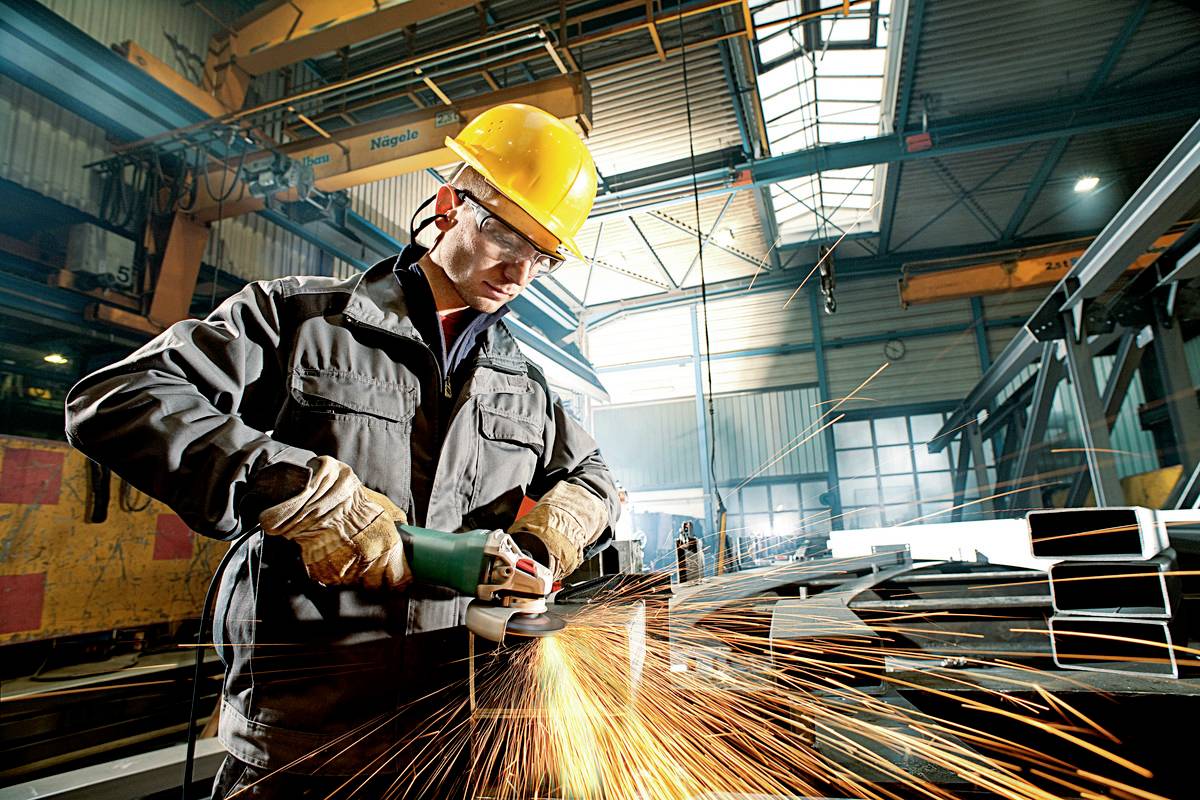 Een fabrieksarbeider met veiligheidshelm en -bril slijpt metaal met een haakse slijper terwijl de vonken rondvliegen.