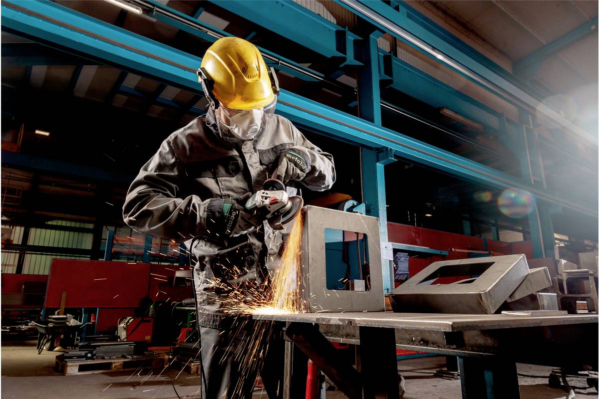 Een arbeider in beschermende uitrusting en helm slijpt metaal met een haakse slijper. Vonken vliegen rond in een industriële omgeving.