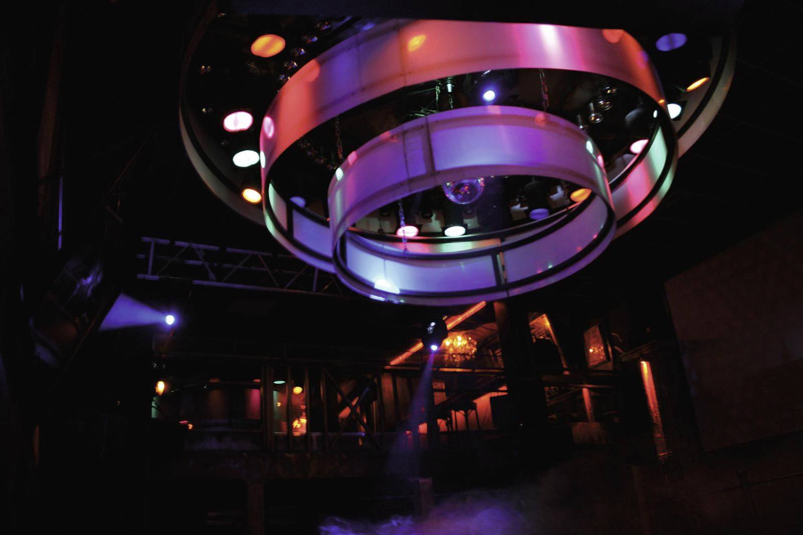 Kleurrijke lampen aan een ronde plafondinstallatie verlichten een donkere clubscène, die een levendige feeststemming oproept.