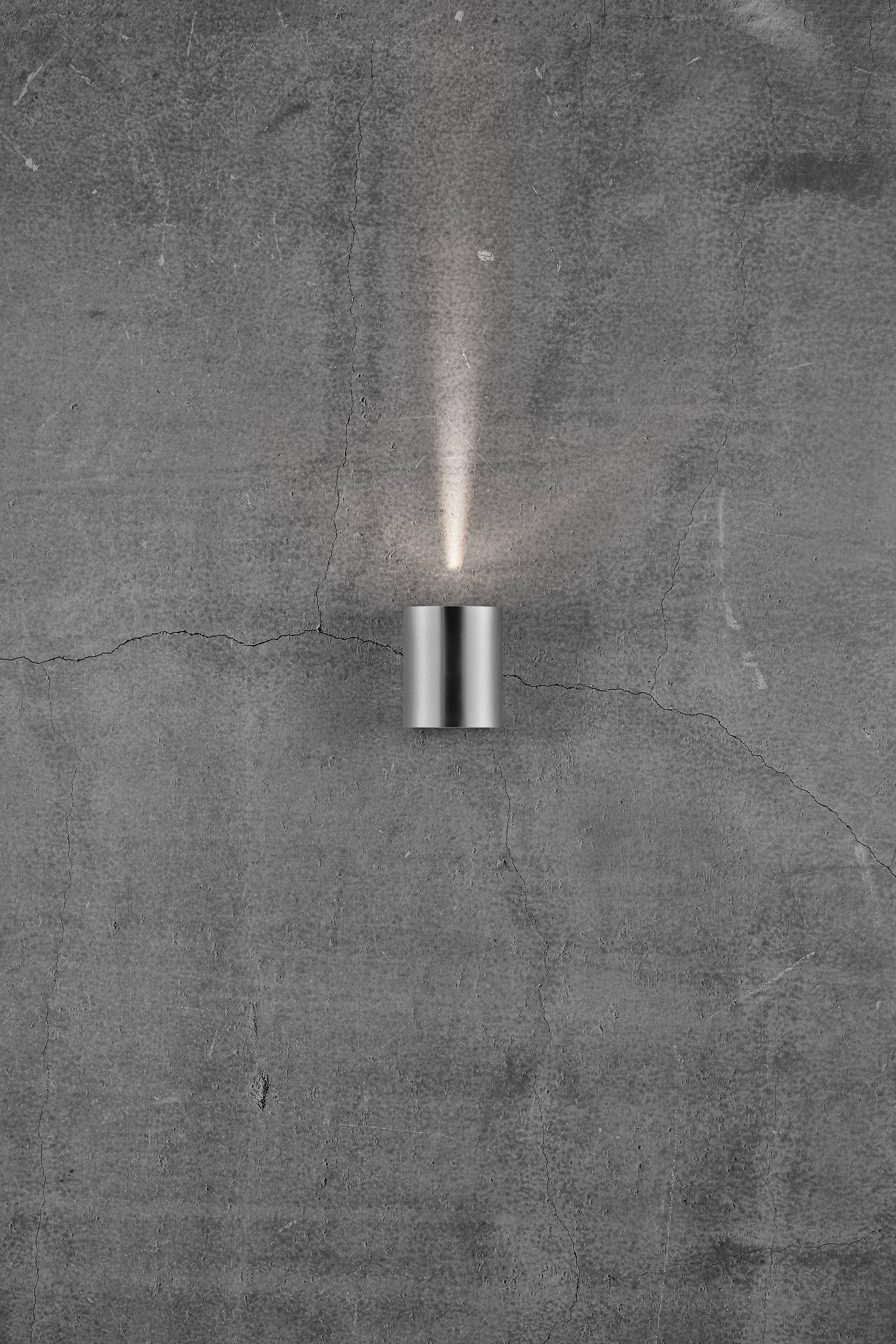 Een moderne wandlamp van metaal met een cilindrisch ontwerp straalt een smalle lichtbundel naar boven op een grijze betonnen muur.