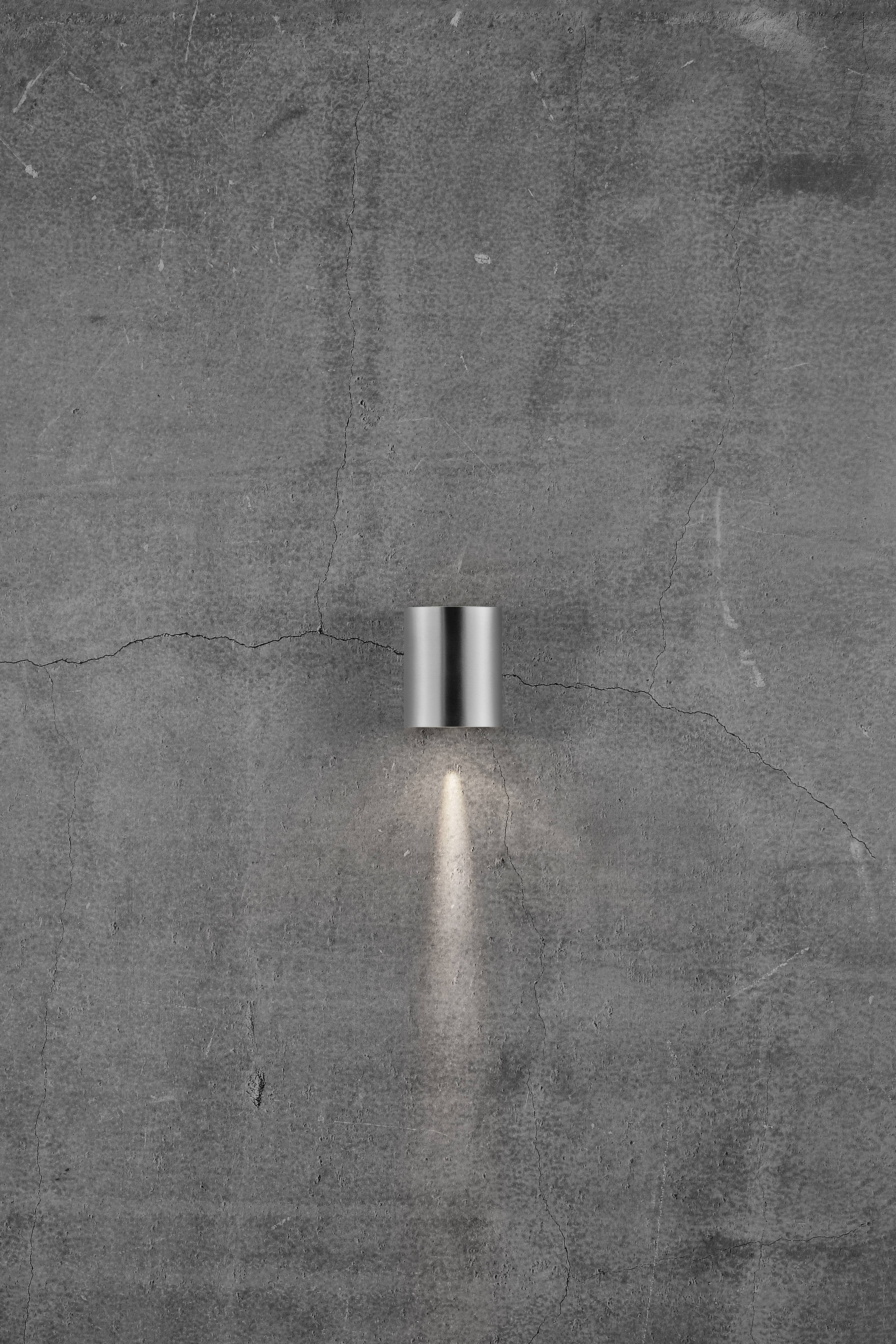 Een moderne, zilveren wandlamp werpt een smalle lichtstraal op een ruwe, grijze betonnen muur.