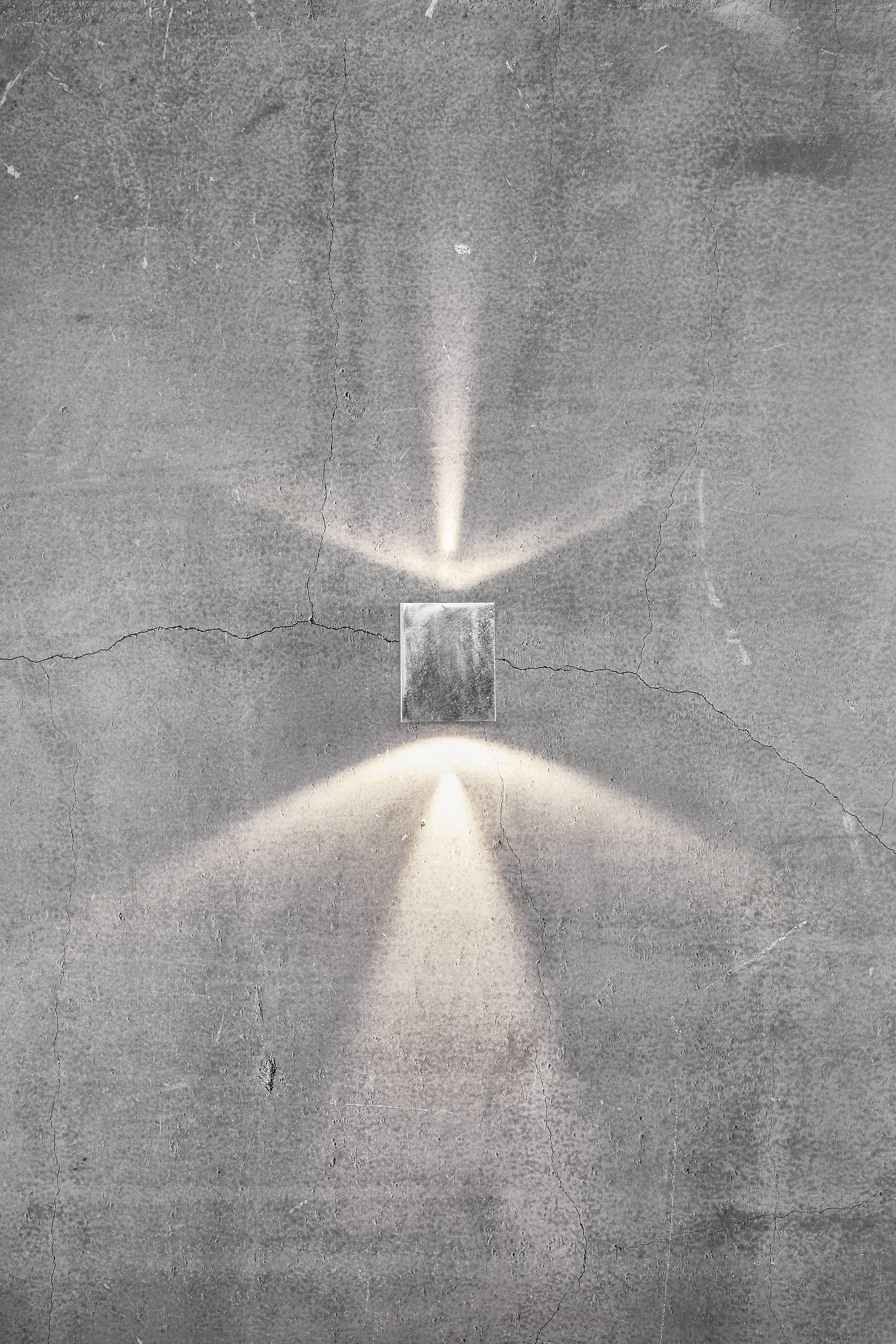 Een enkele wandlamp straalt lichtstralen naar boven en beneden op een grijze, ruwe betonwand.