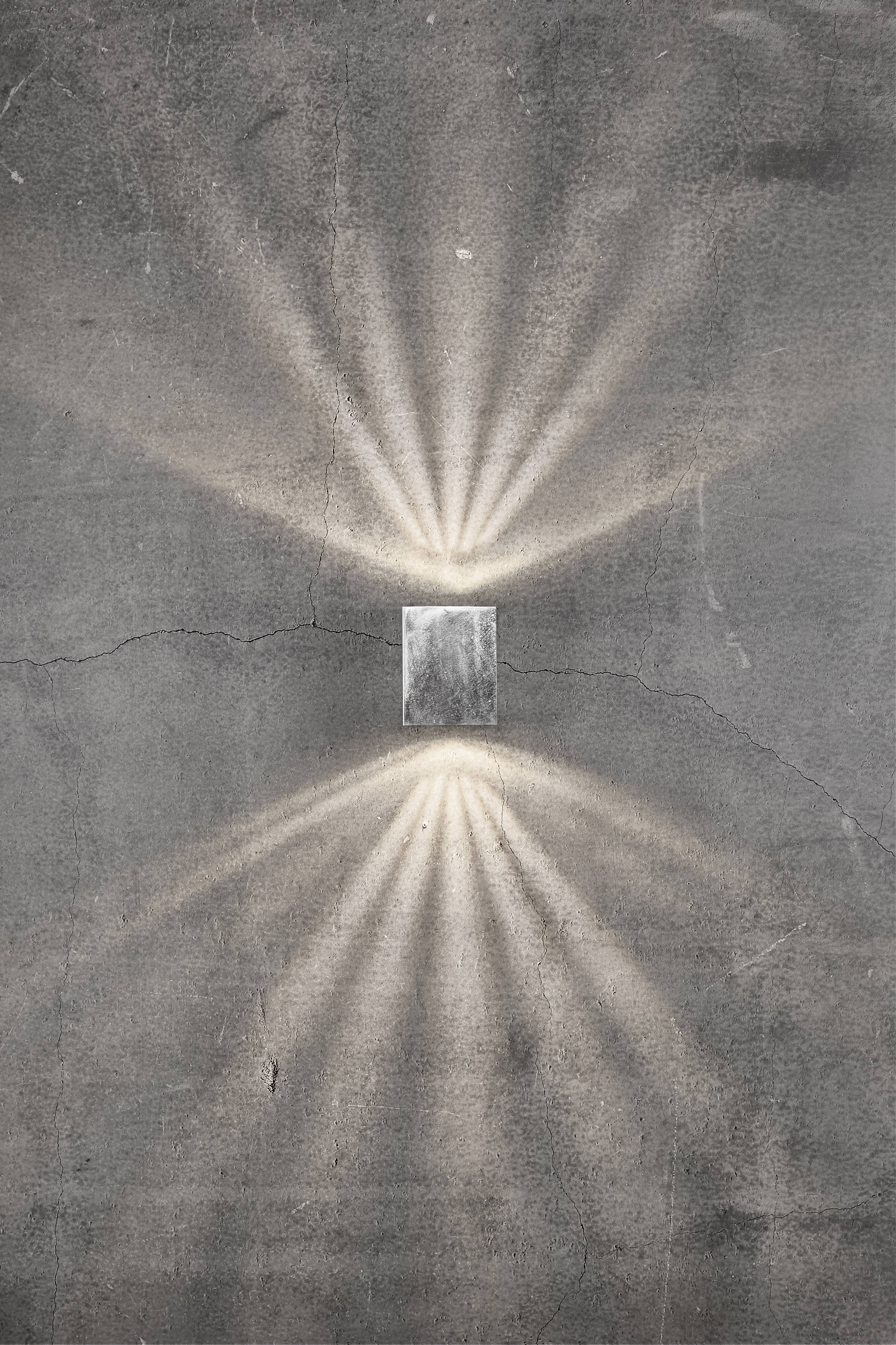 Een rechthoekige wandlamp op een grijze betonnen muur straalt lichtstralen naar boven en beneden uit en creëert een symmetrisch patroon.