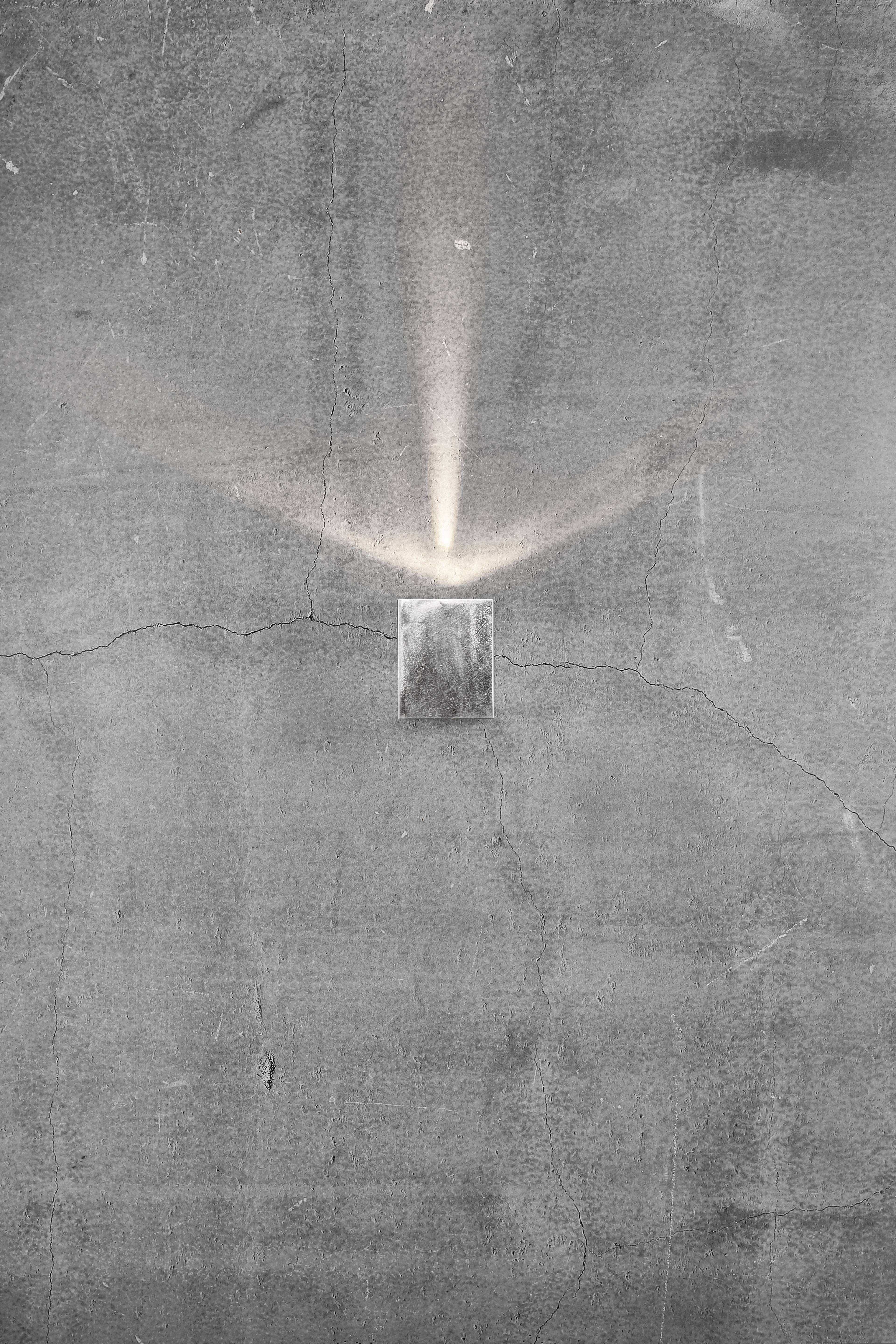 Een kleine lichtstraal schijnt uit een rechthoekig metalen voorwerp tegen een grijze, gebarsten betonnen muur.