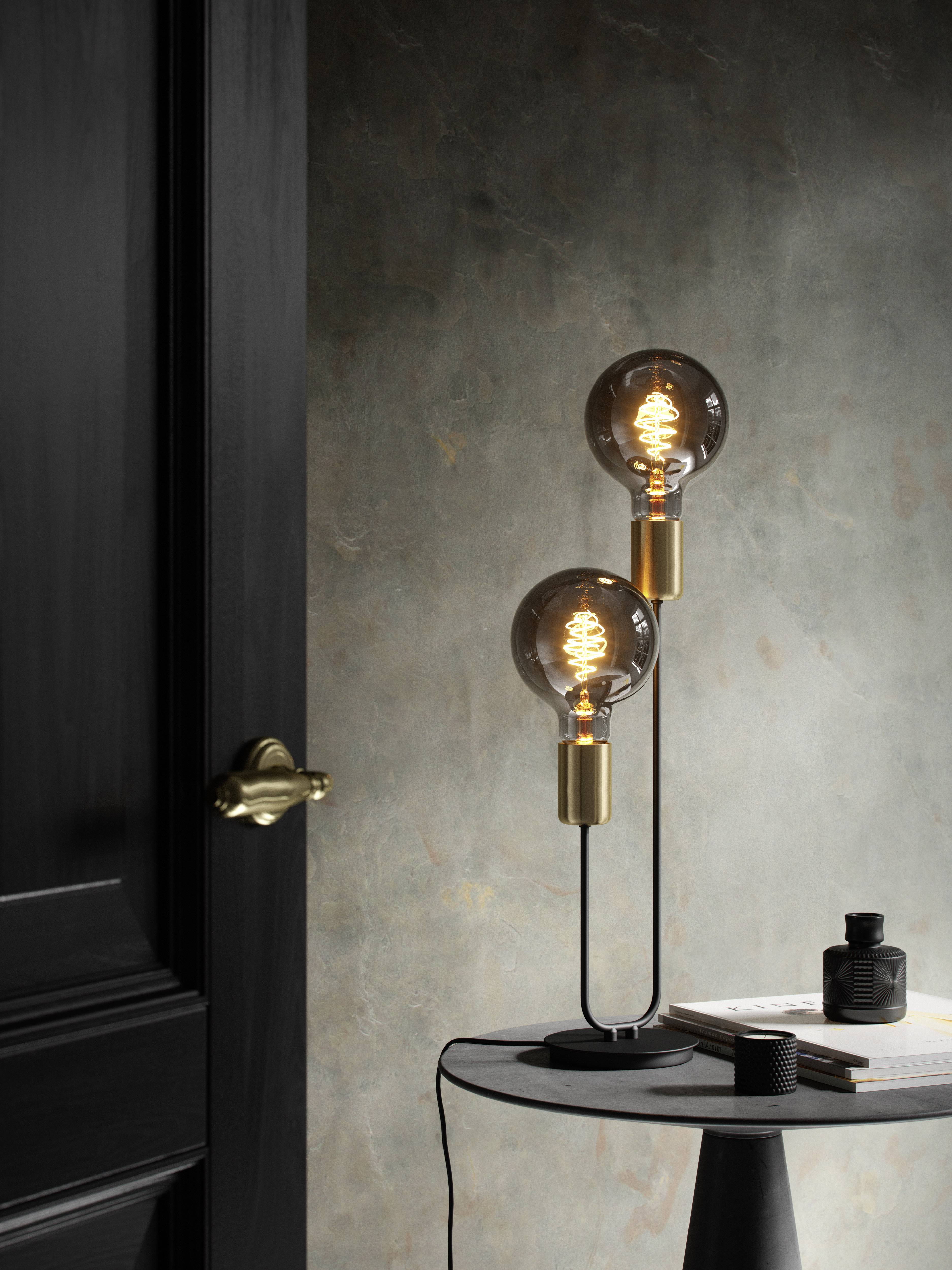 Staande lamp met twee lichtgevende gloeilampen op een kleine tafel naast een zwarte deur, minimalistisch interieur.