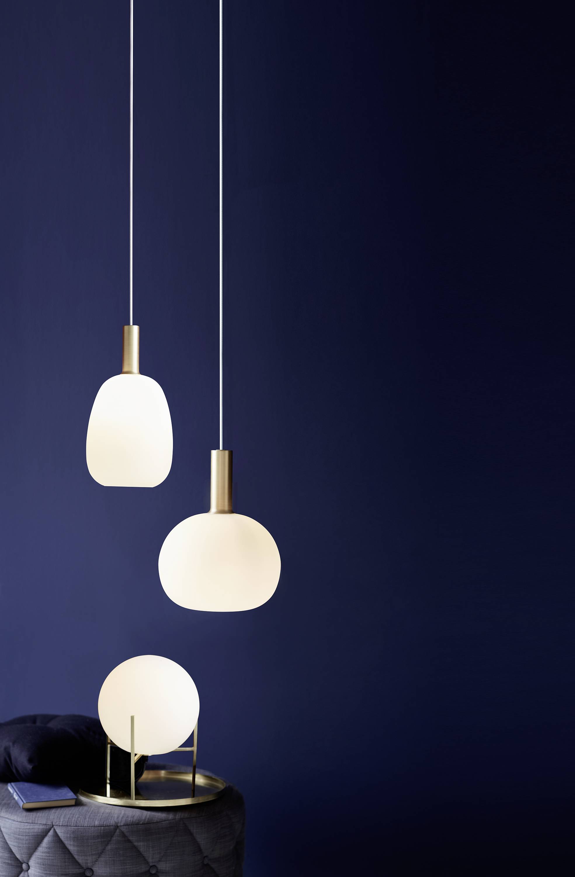 Drie moderne witte hanglampen voor een donkerblauwe achtergrond, daaronder een kleine tafel met kussens en een ronde tafellamp.