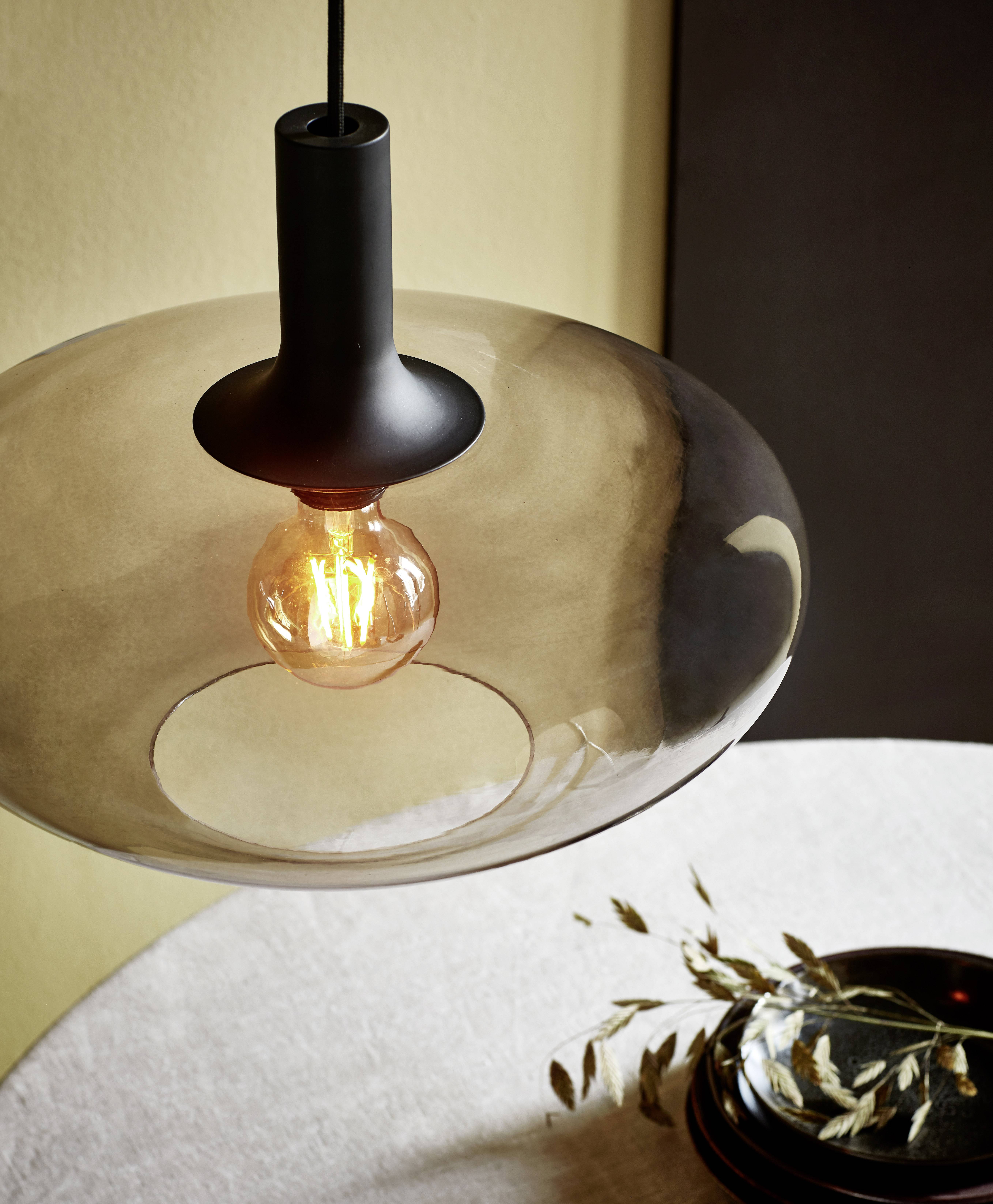 Een moderne hanglamp met een glazen kap en zichtbare gloeilamp hangt boven een tafel met een decoratieve tak en een schaal.
