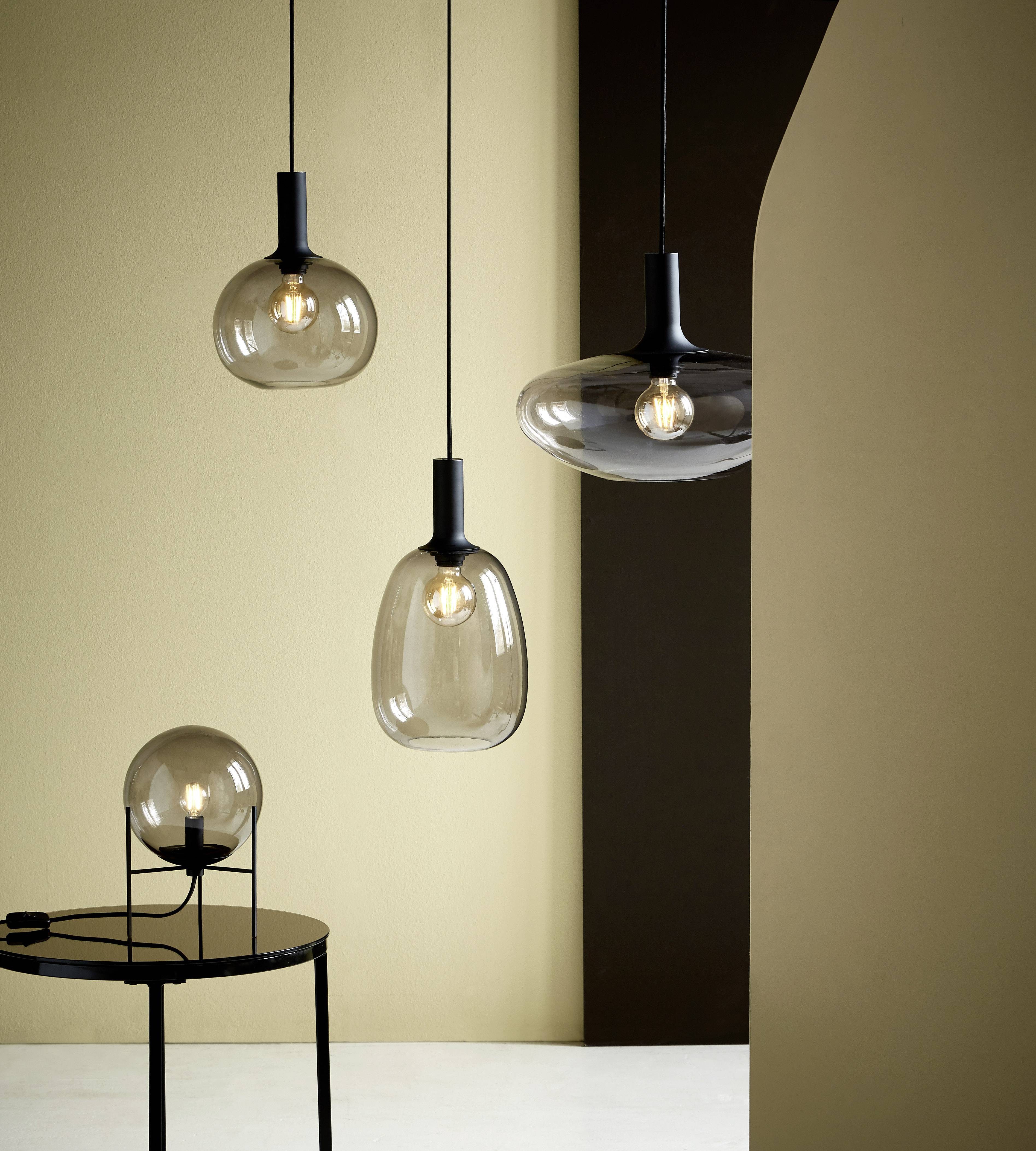 Drie moderne hanglampen met verschillende glazen bollen hangen aan het plafond, naast een tafelamp die op een zwarte tafel staat.