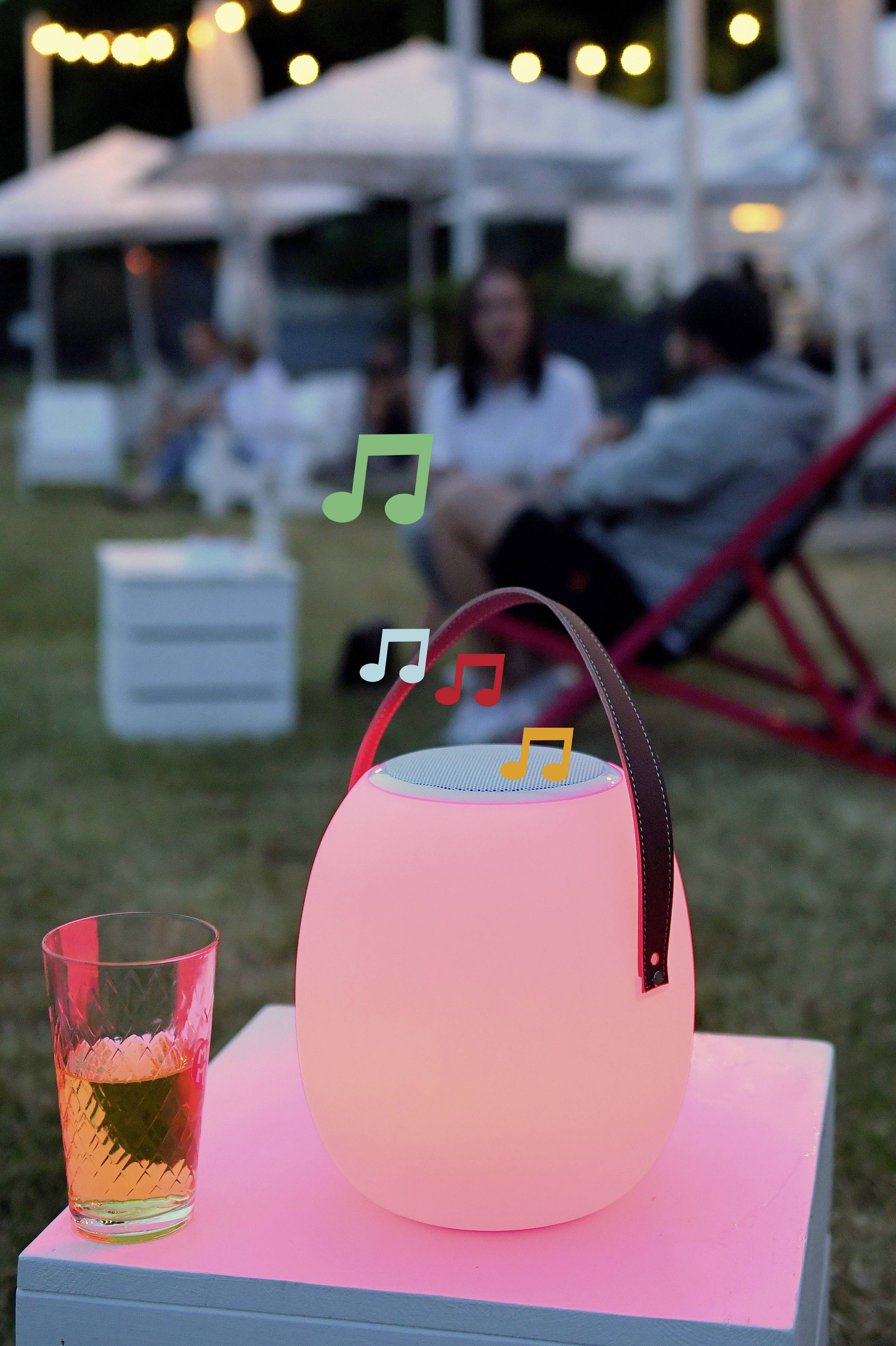 Een kleurrijke, stralende lamp op een tafel met muzieksymbolen. Op de achtergrond zitten personen onscherp op een grasveld.