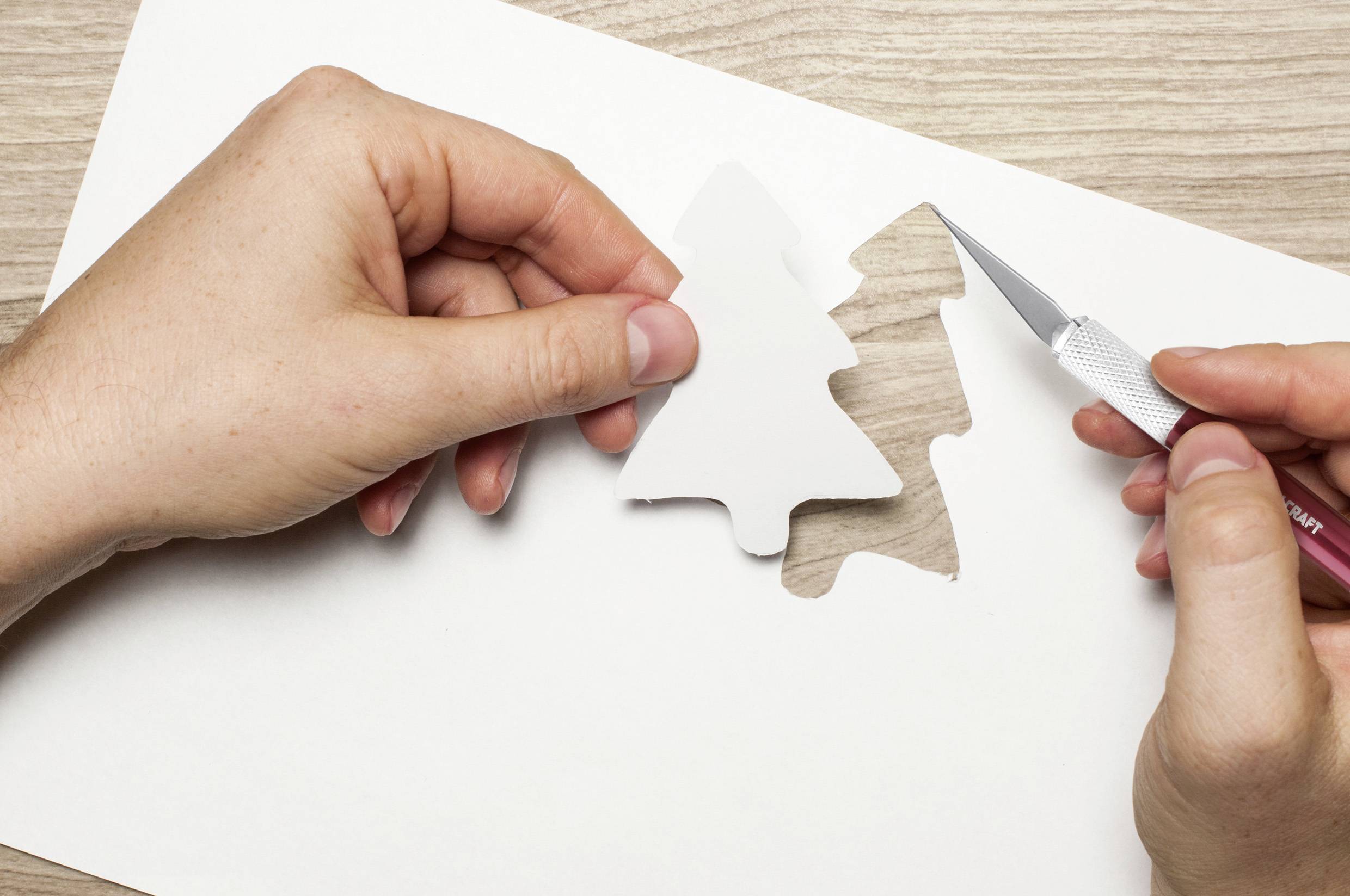 Een hand snijdt met een scalpel een dennenboomvorm uit wit papier, terwijl de andere hand de vorm vasthoudt.