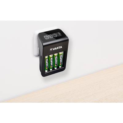 Een batterijlader van Varta steekt in een stopcontact aan de muur en laadt vier AA-batterijen. Hij bevindt zich in de buurt van een houten tafelblad.