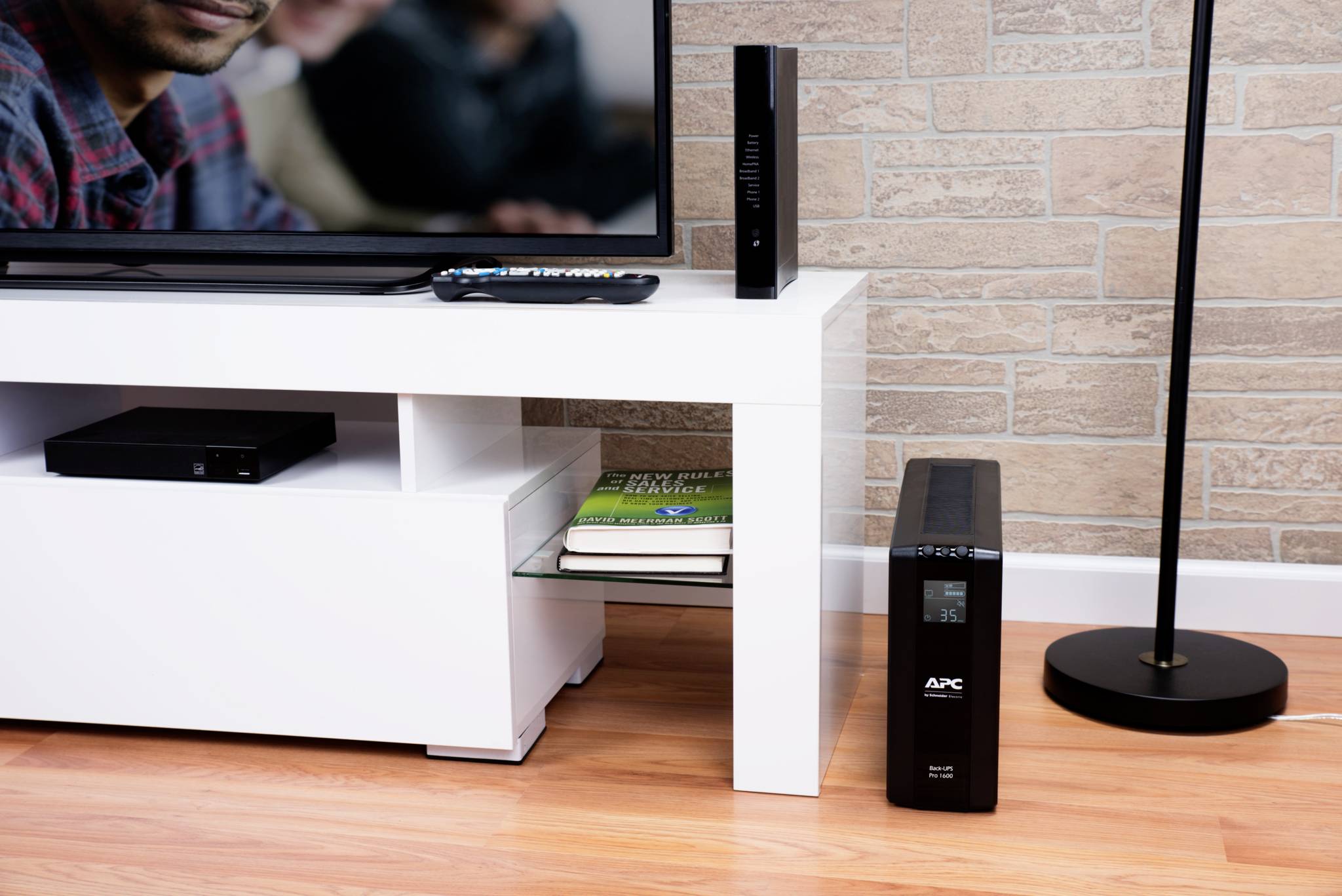 Een woonkamer met een witte tv-kast, waarop een televisie, een afstandsbediening en een router staan. Ernaast een APC-UPS-apparaat.